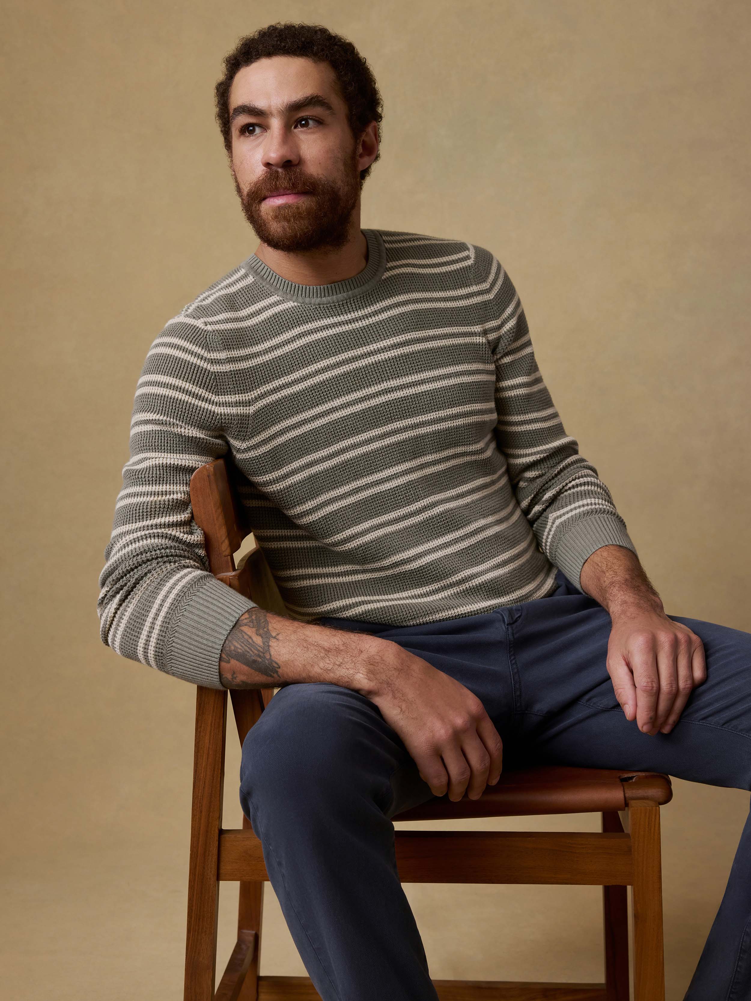 Man sitting on a chair wearing a green striped sweater and navy pants against a beige background