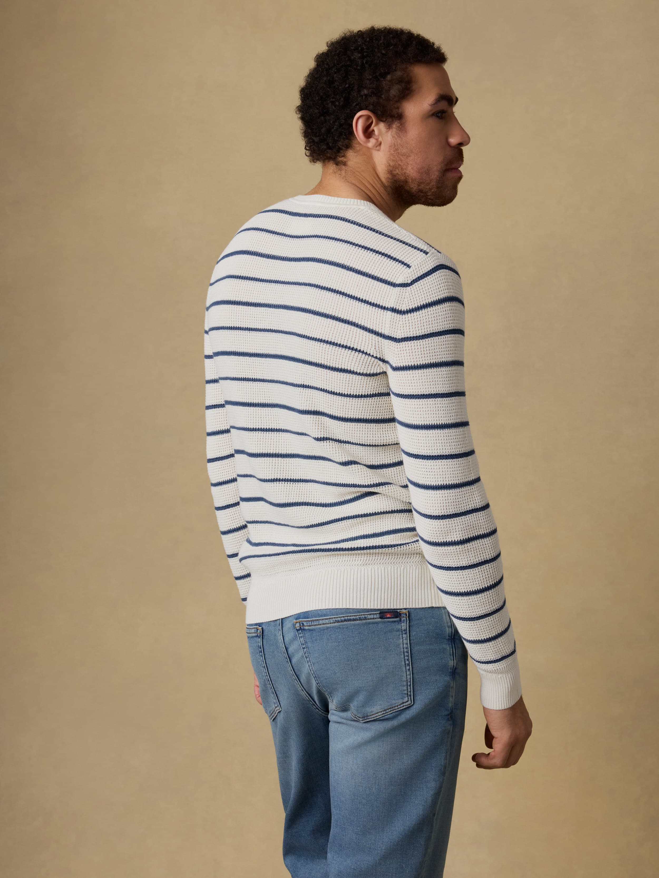 Back view of a man wearing a white and blue striped crewneck sweater and blue jeans against a beige background