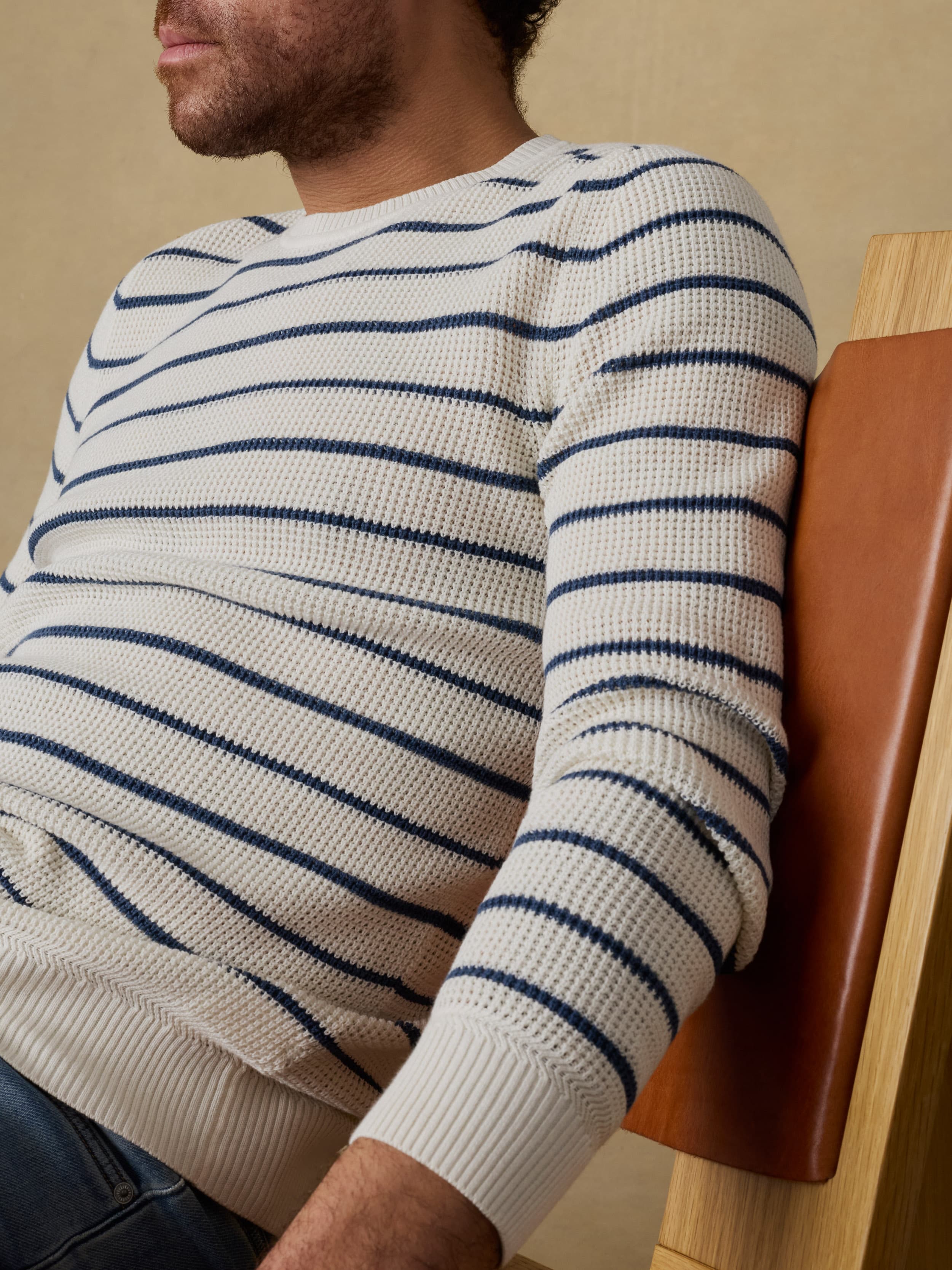 Man sitting on a wooden chair wearing a white and blue striped crewneck sweater and blue jeans against a beige background