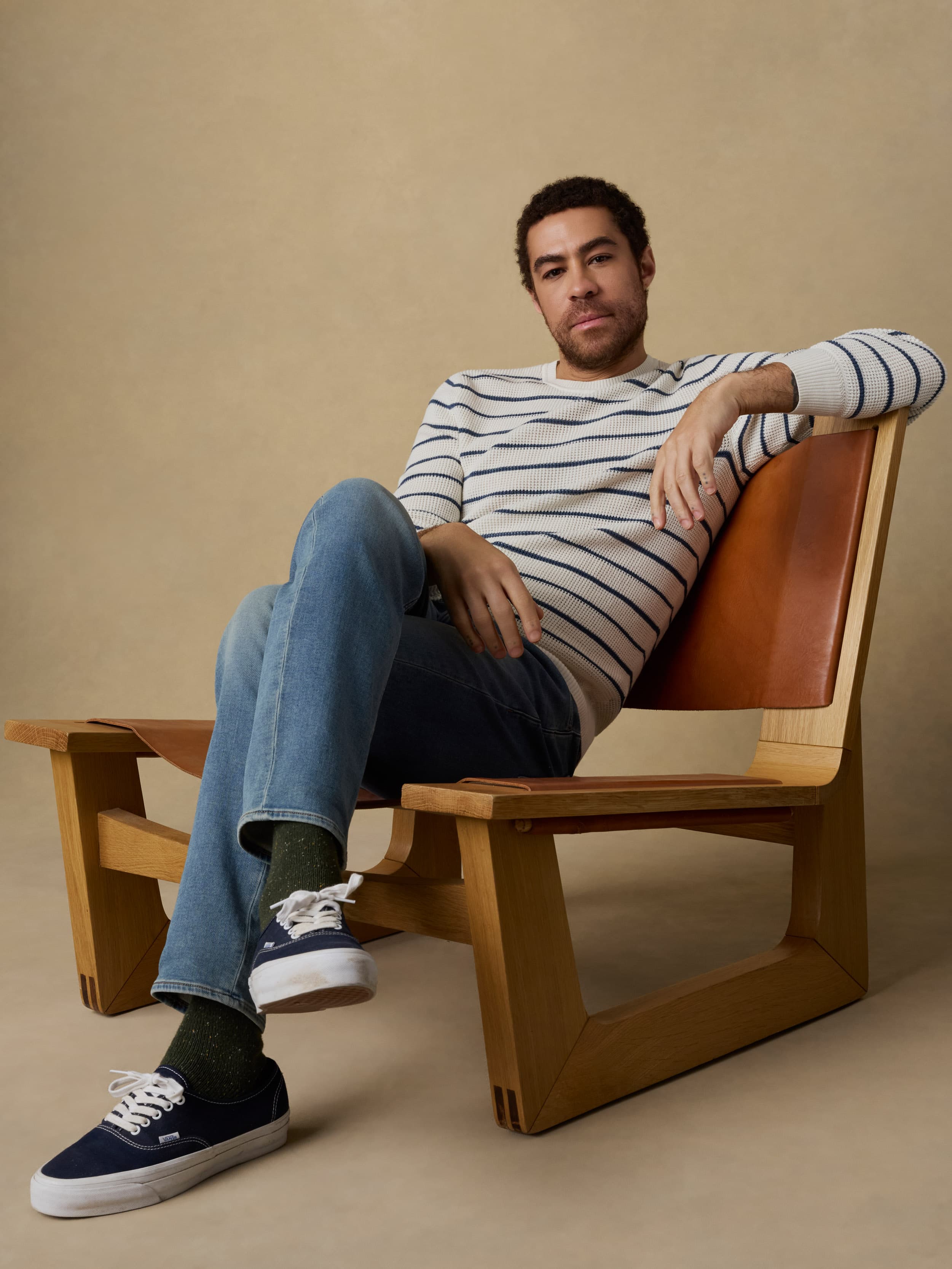 Man sitting on a wooden chair wearing a white and blue striped crewneck sweater and blue jeans against a beige background