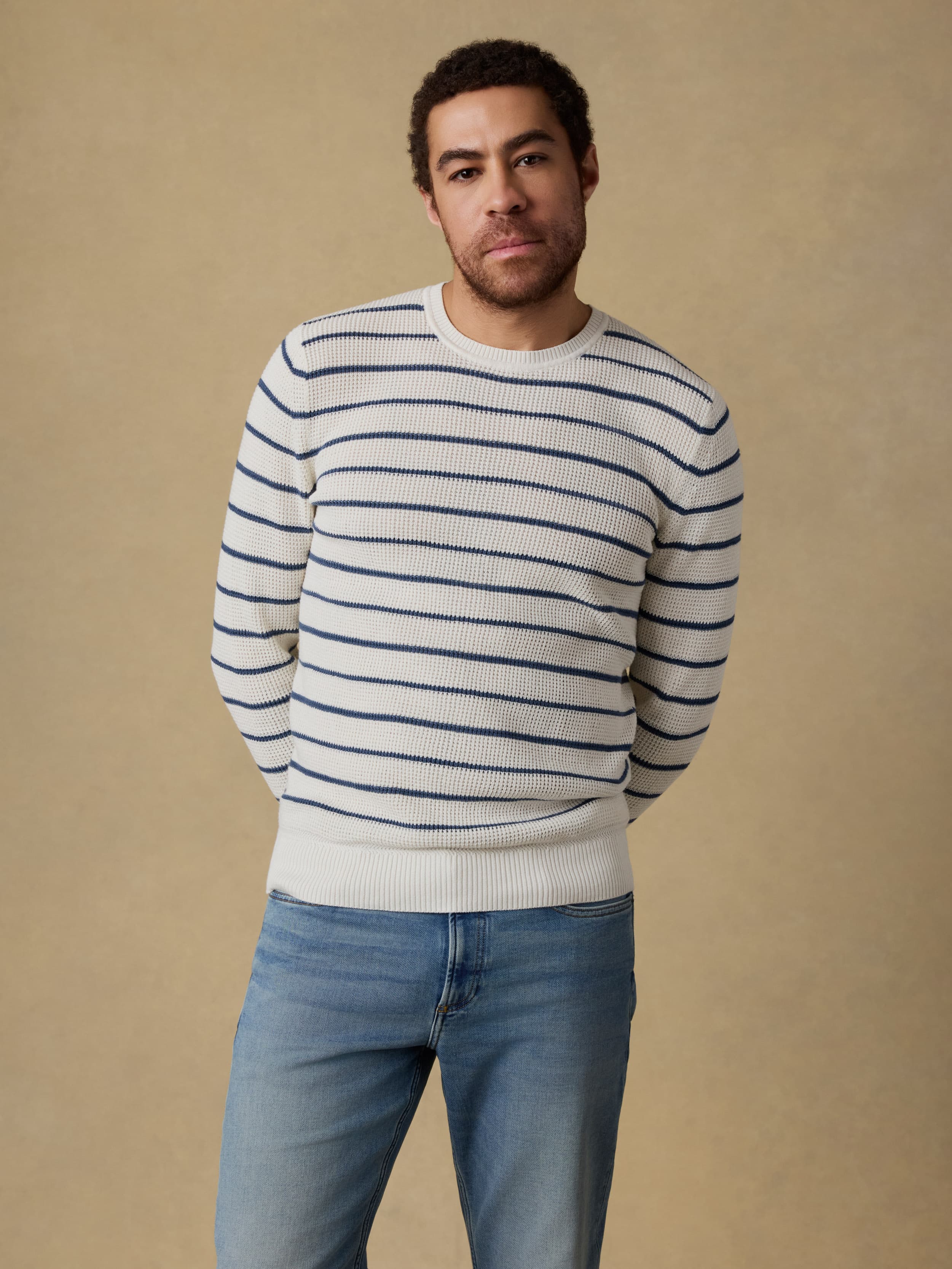 Man wearing a white and blue striped crewneck sweater and blue jeans against a beige background