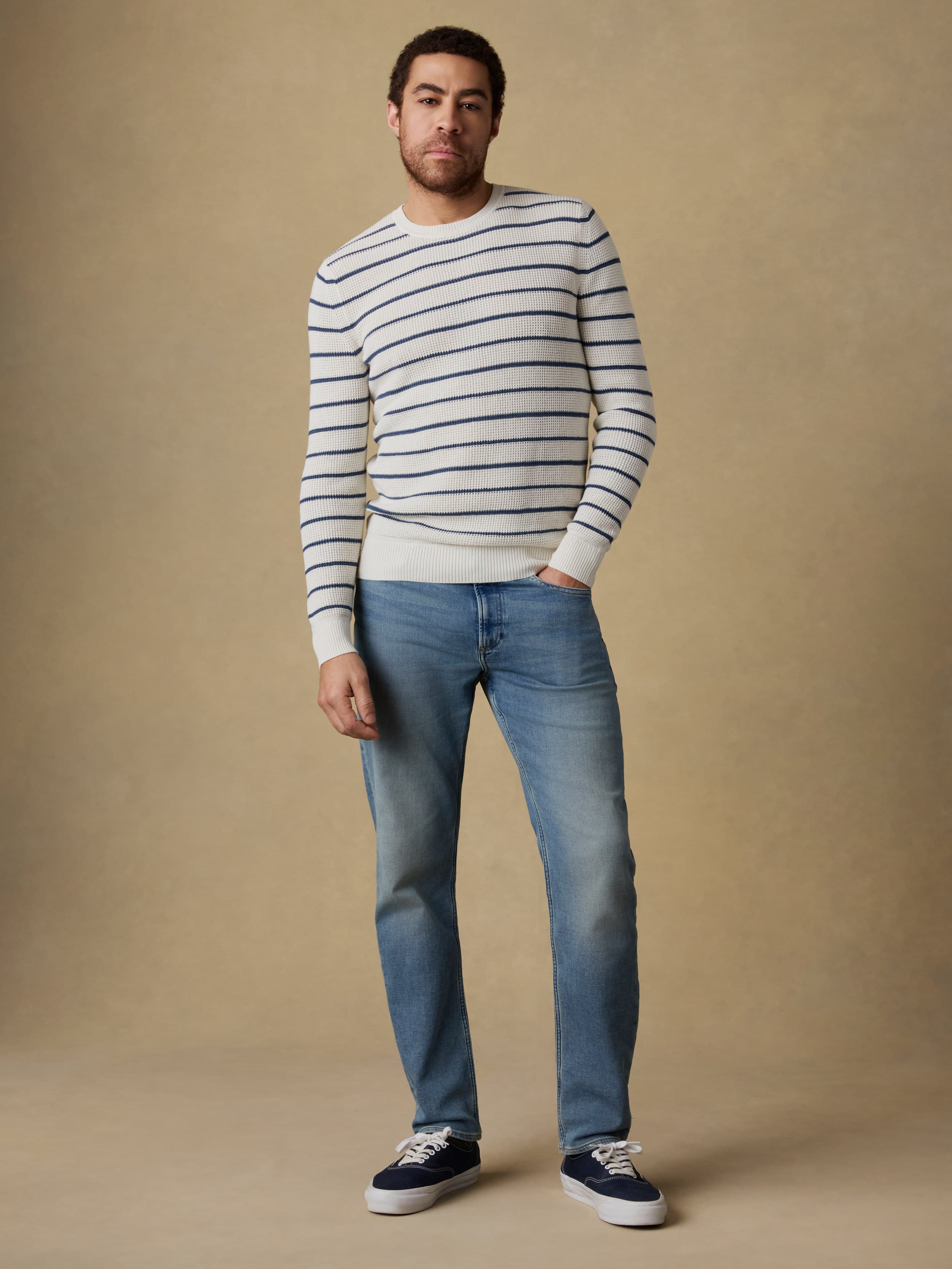 Man wearing a white and blue striped crewneck sweater and blue jeans against a beige background