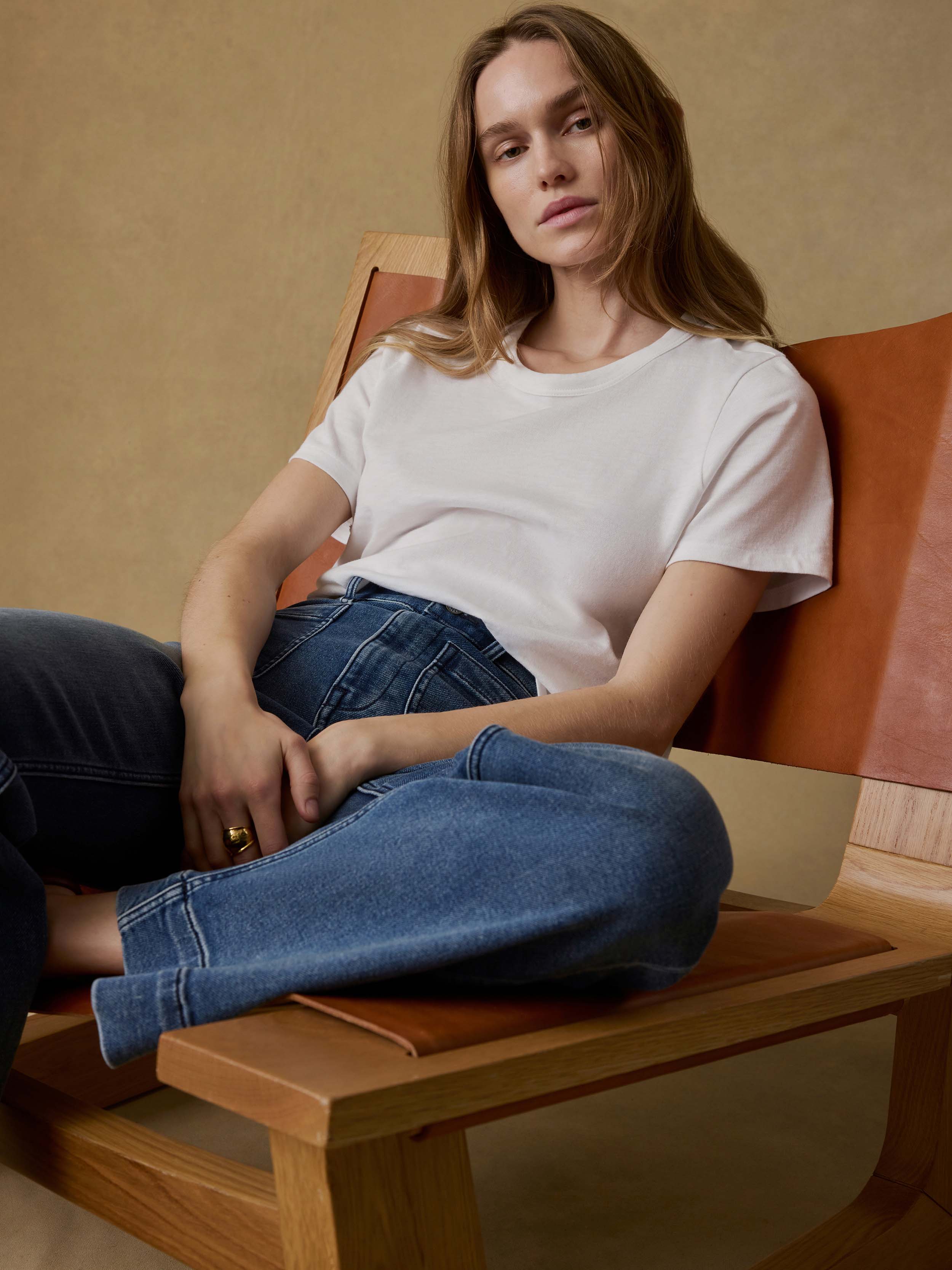 Woman sitting on a wooden chair wearing a white t-shirt and blue jeans against a beige background.