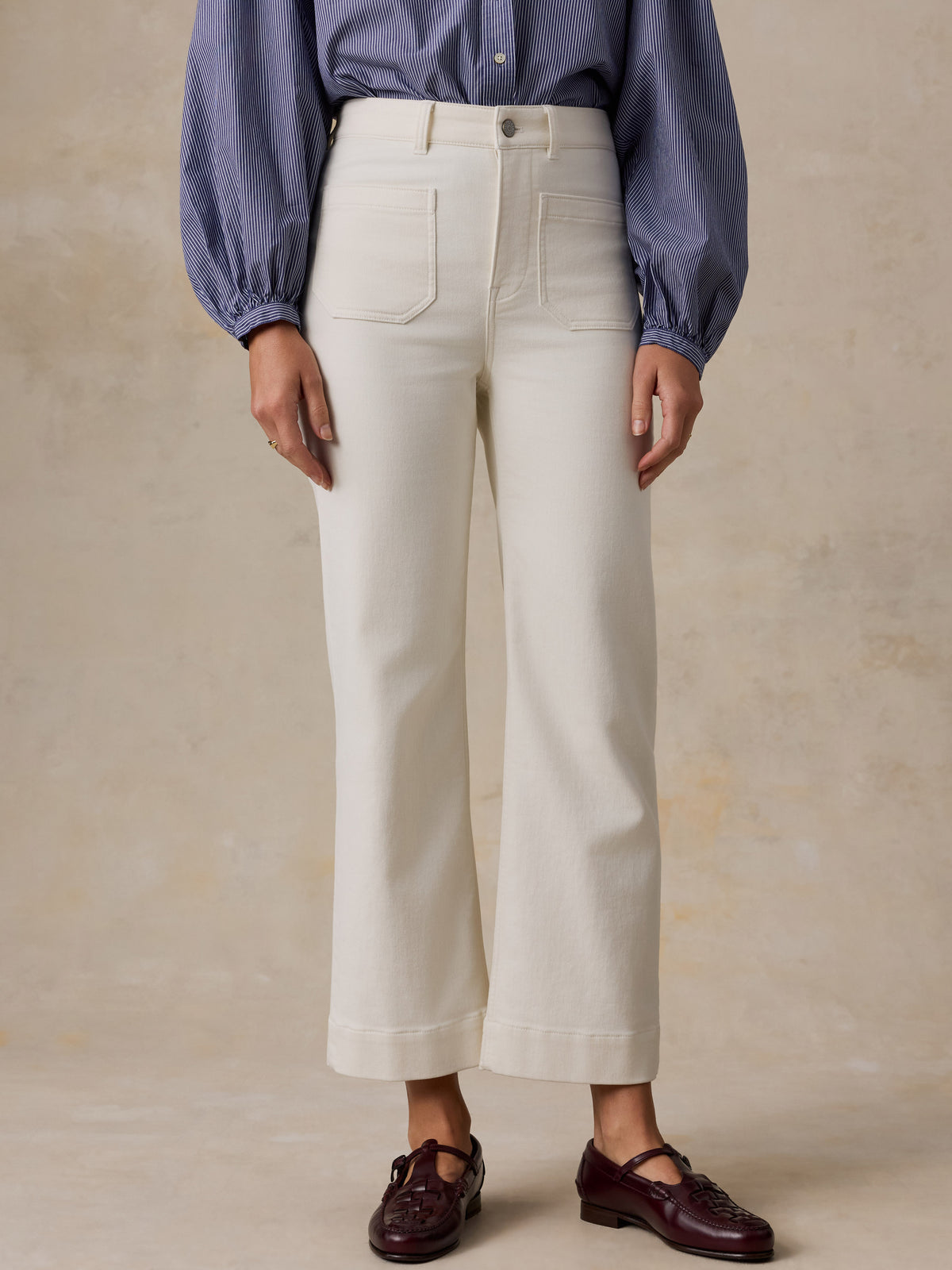 A model is shown from the waist down wearing a pair of off-white cropped wide-leg jeans, a blue striped long-sleeve blouse, and dark burgundy Mary Jane shoes, set against a textured beige studio background.