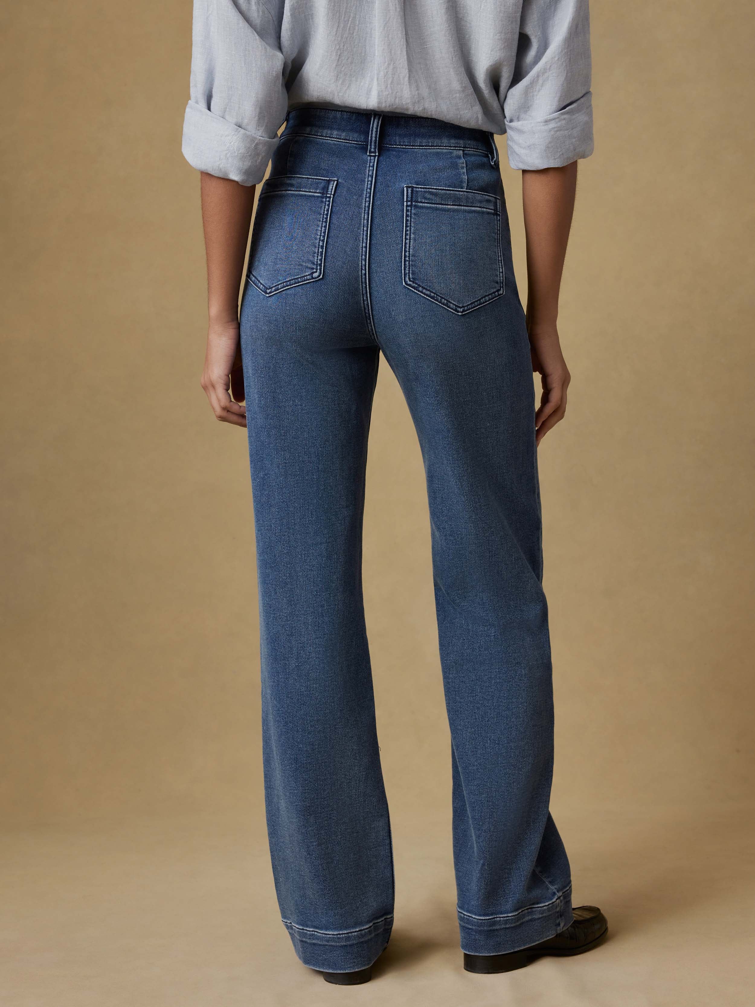 Back view of a woman wearing a light blue button up shirt and dark blue jeans against a beige background