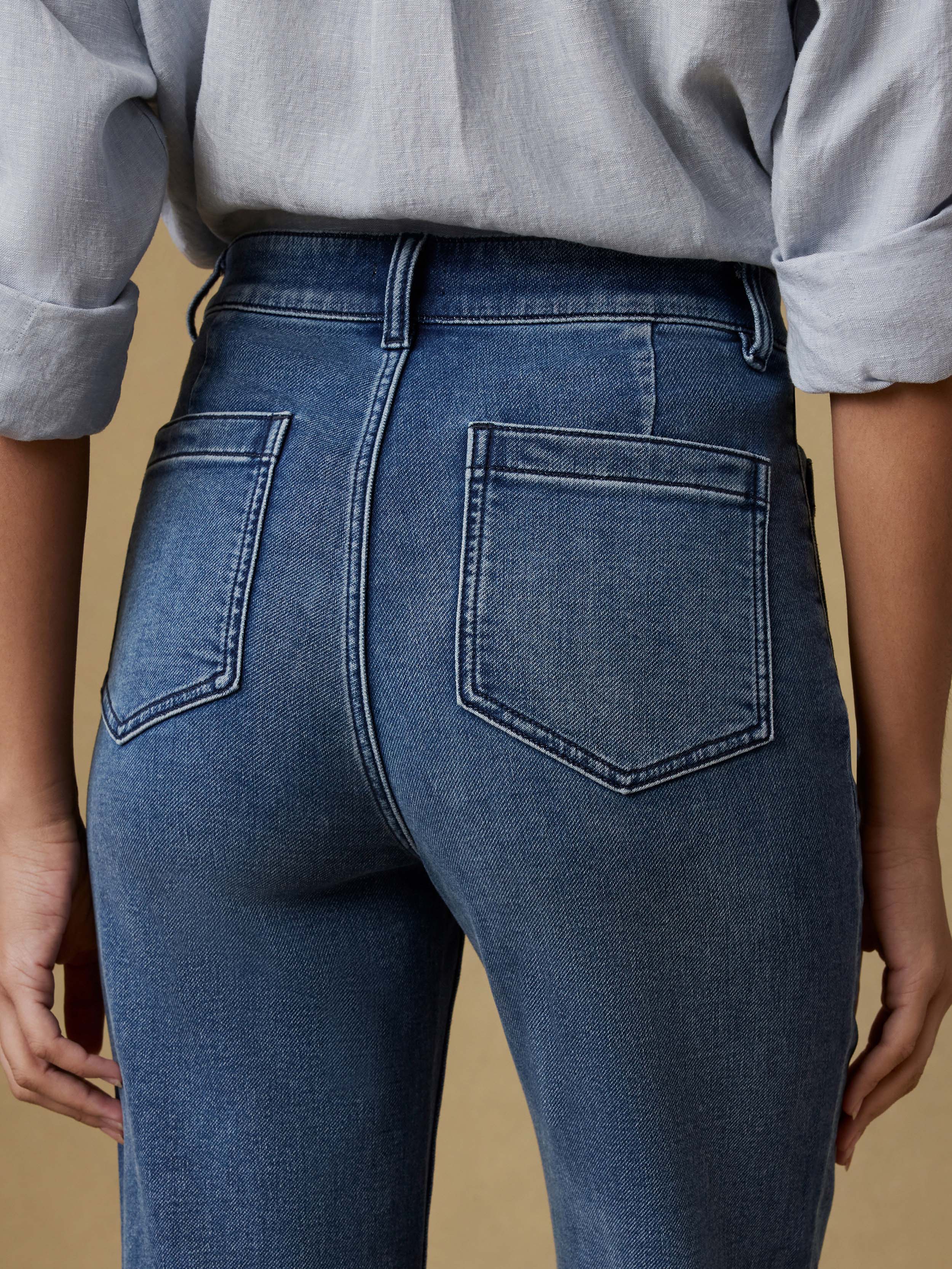 Detail back view of a woman wearing a light blue button up shirt and dark blue jeans against a beige background