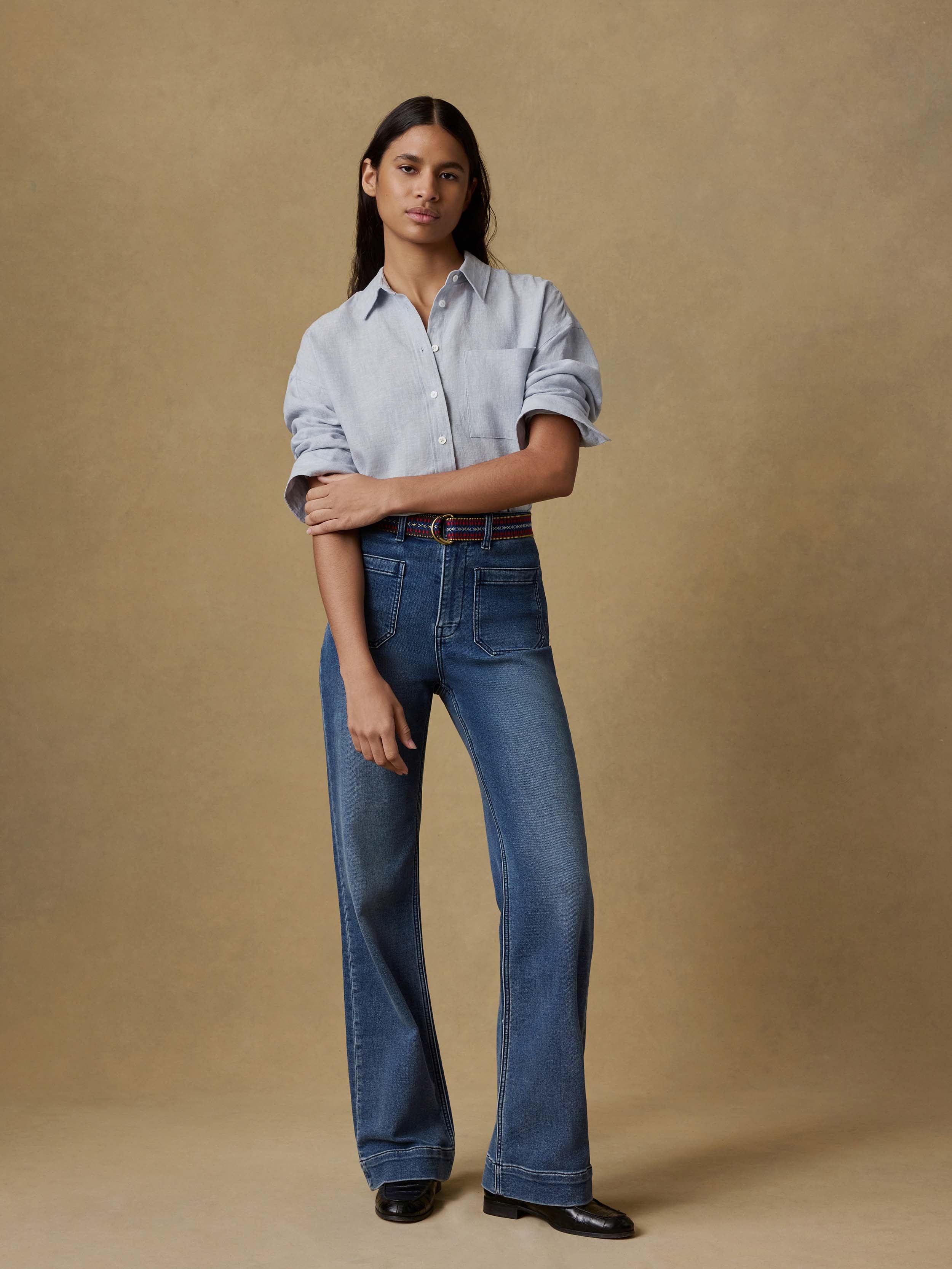 Woman wearing a light blue button up shirt and dark blue jeans against a beige background