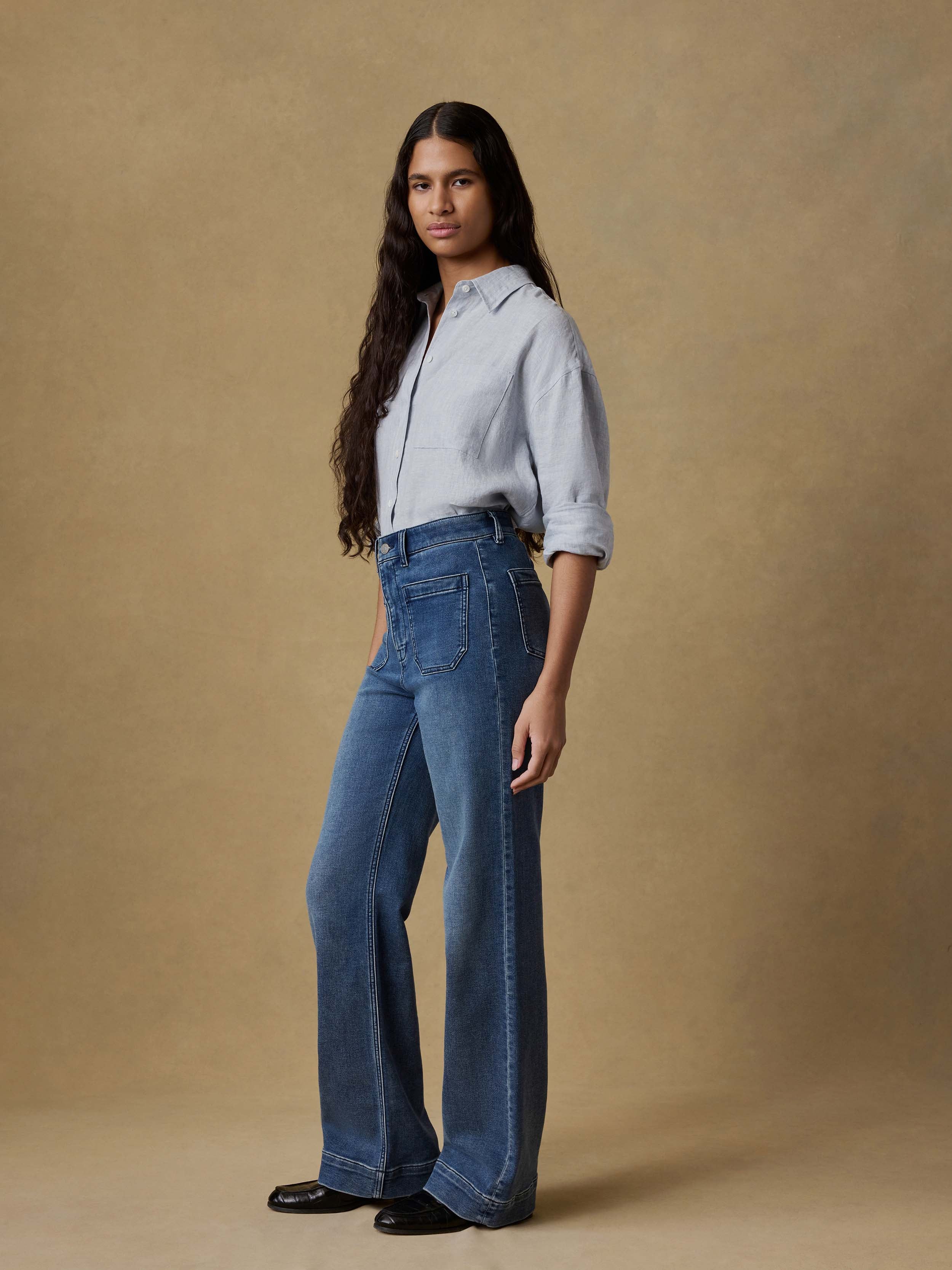 Woman wearing a light blue button up shirt and dark blue jeans against a beige background