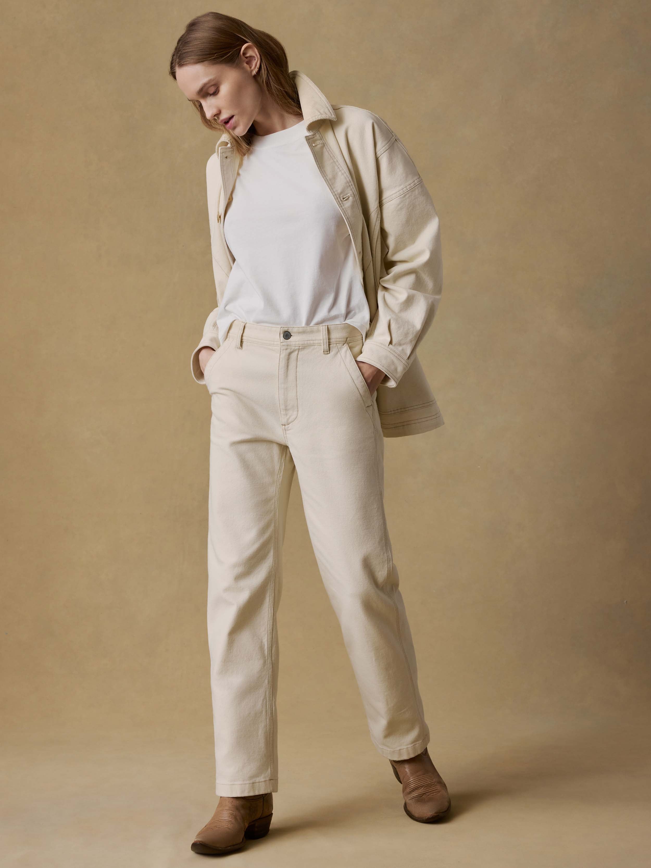 Woman wearing a beige coat, white t-shirt, and beige pants against a beige background