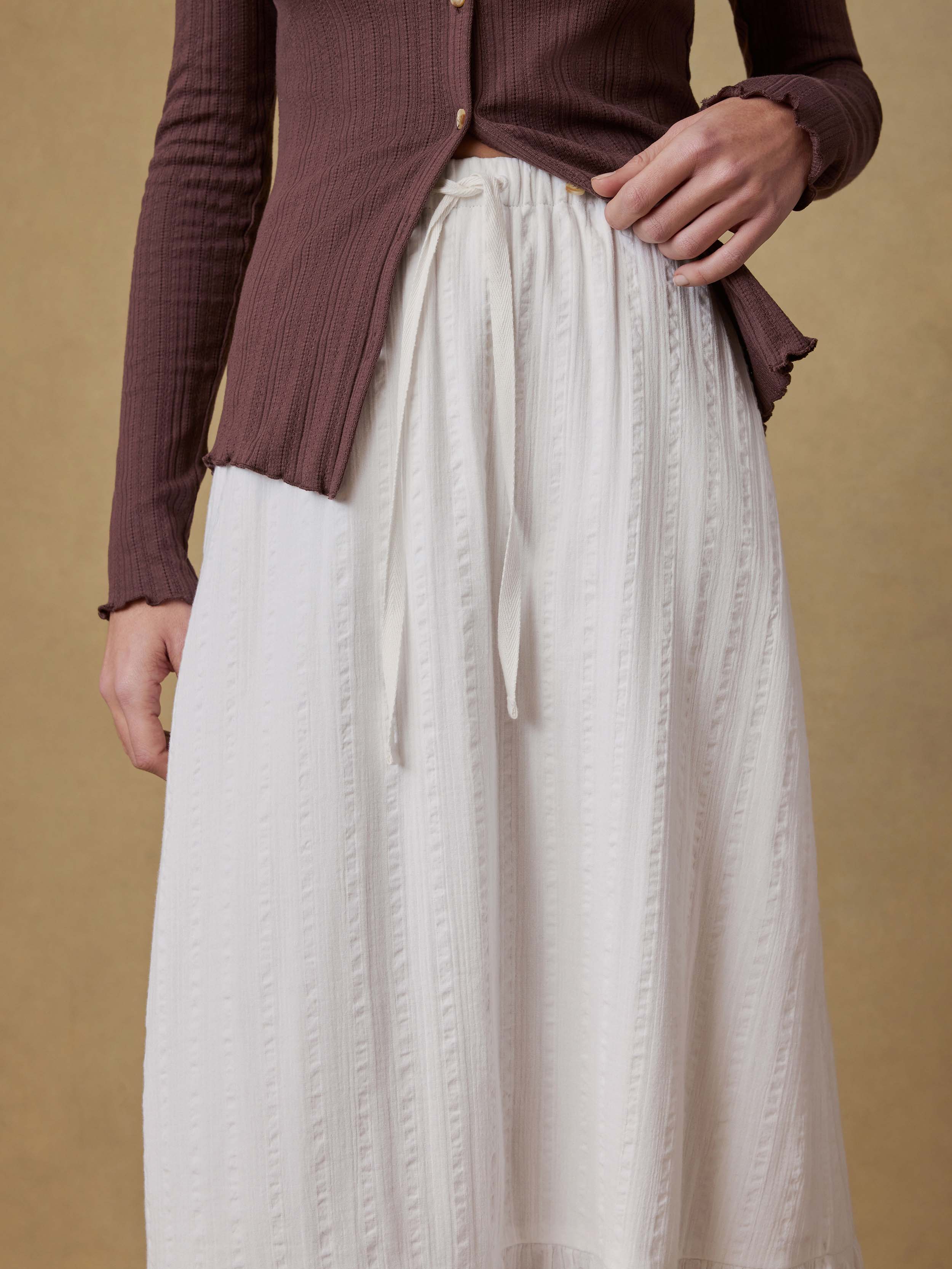 Woman wearing a brown cardigan and white skirt against a beige background