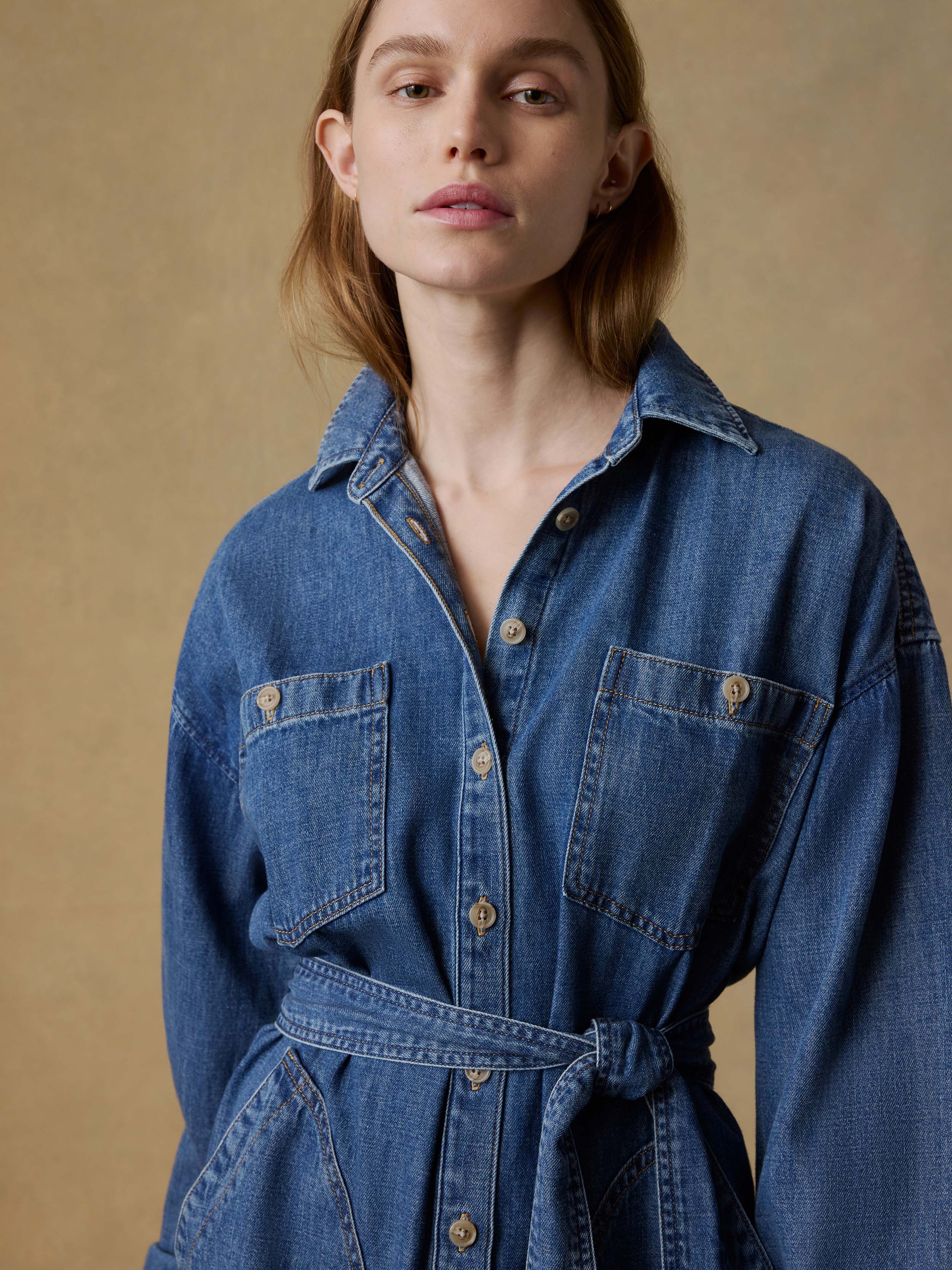Woman wearing a long blue denim dress with a belt on a beige background