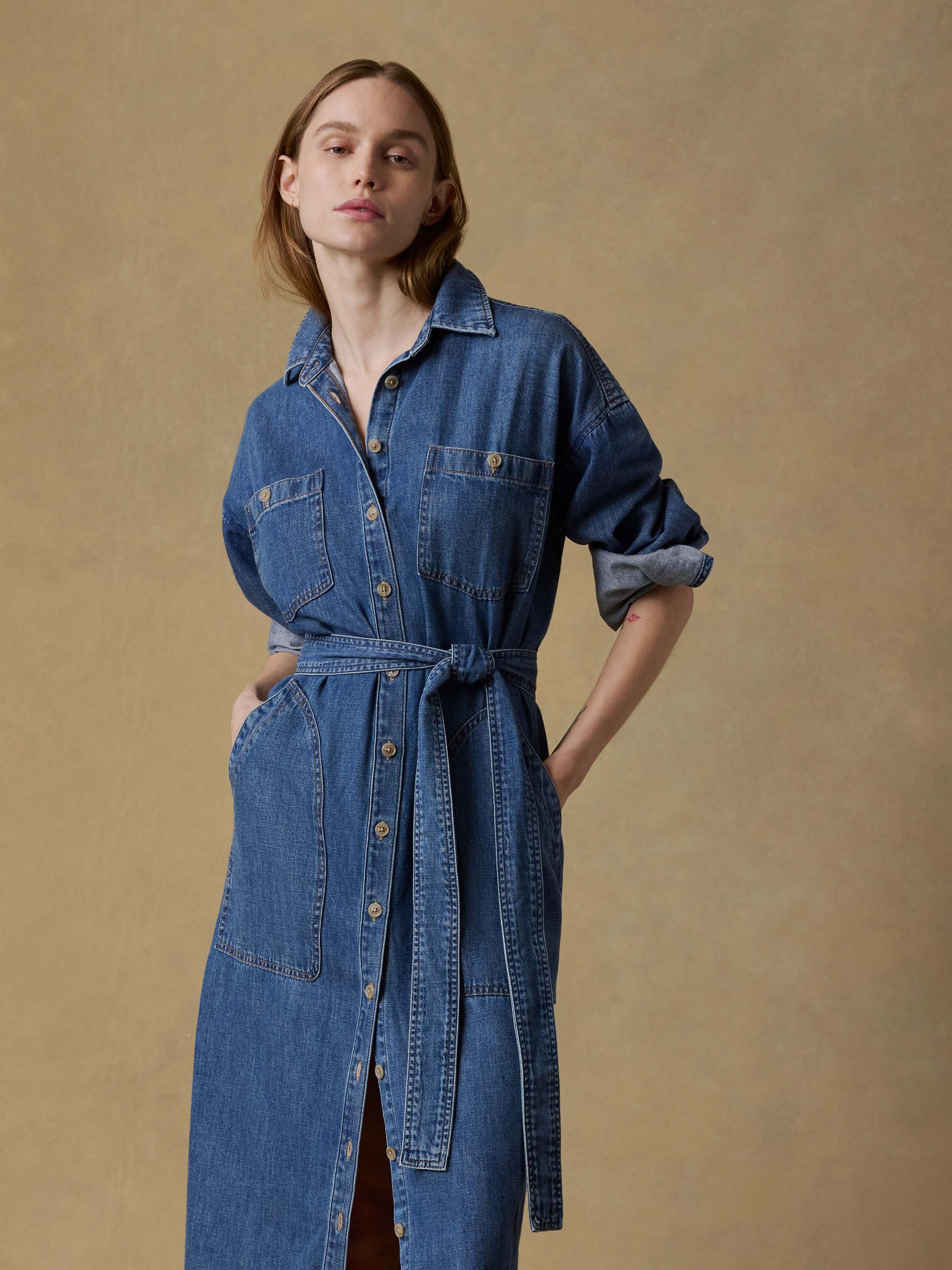 Woman wearing a long blue denim dress with a belt on a beige background