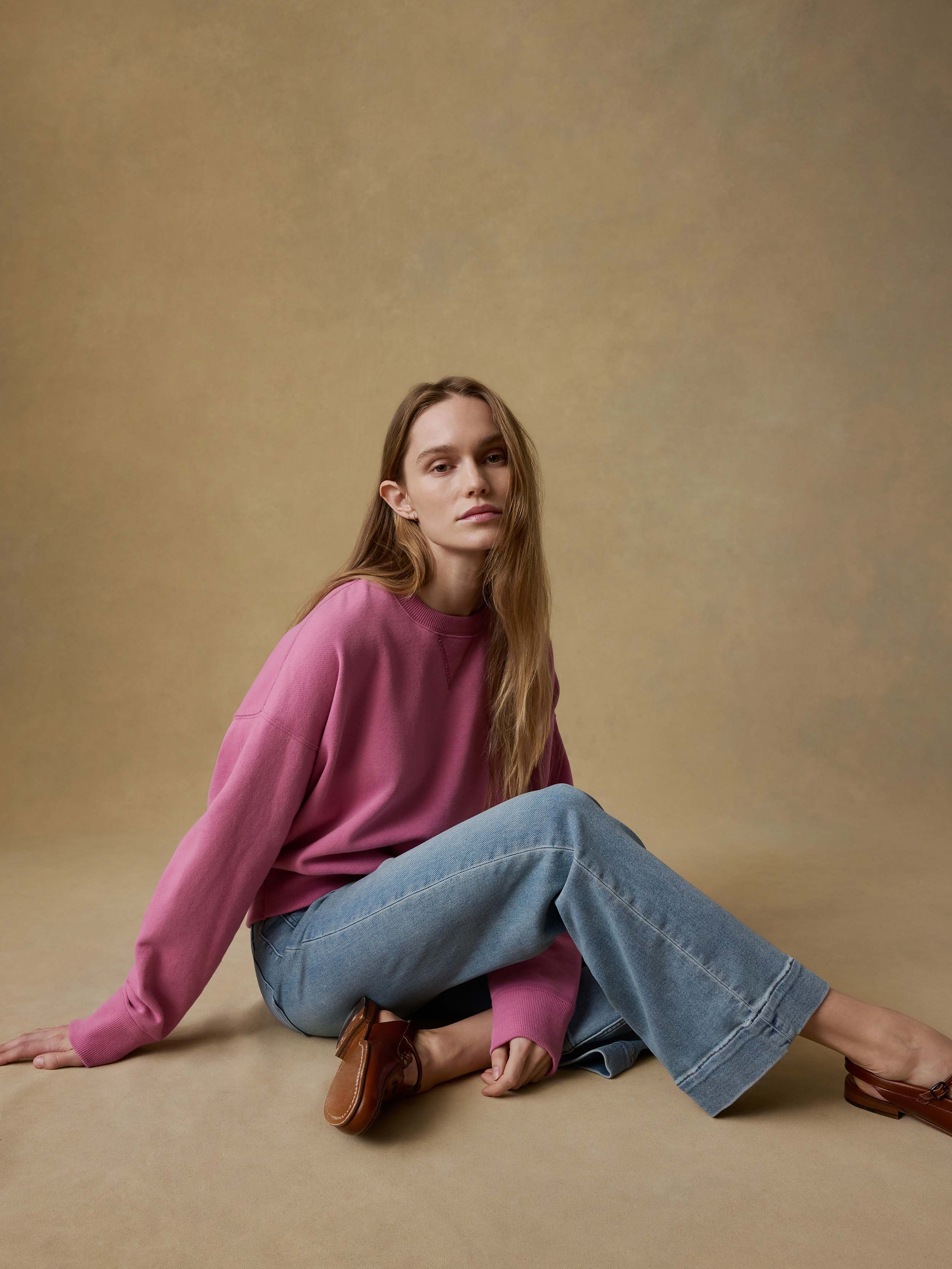 Woman wearing a pink sweatshirt and blue jeans sitting on a beige floor against a beige background.