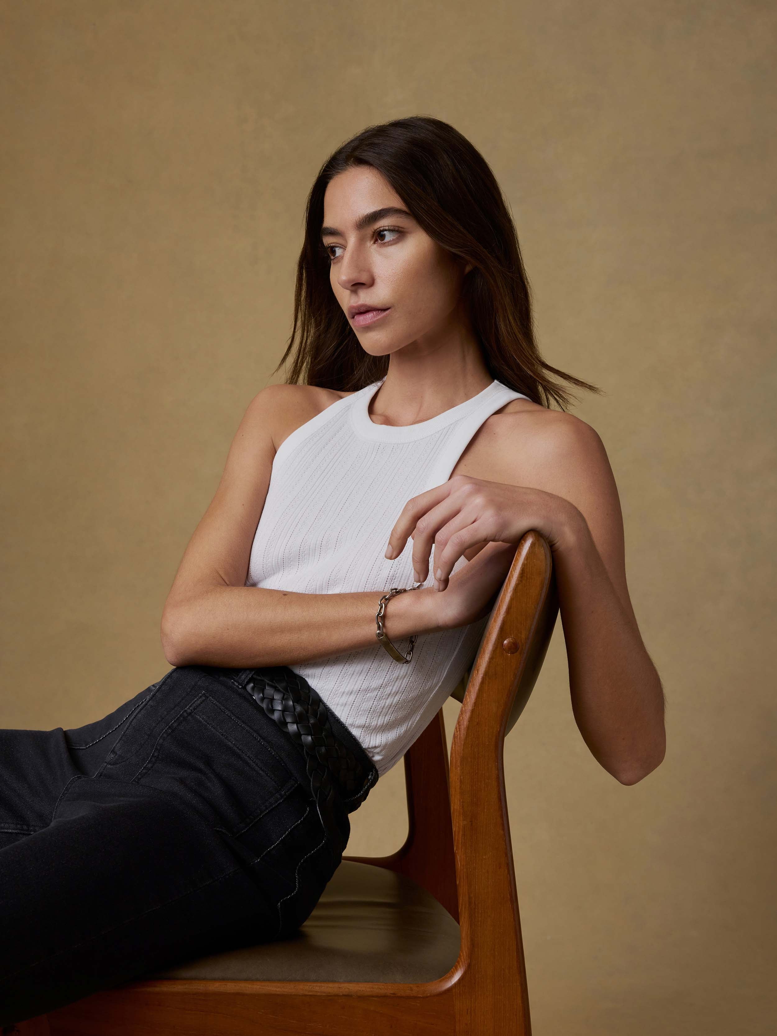 Woman sitting on a wooden chair wearing a white tank top and black jeans against a beige background