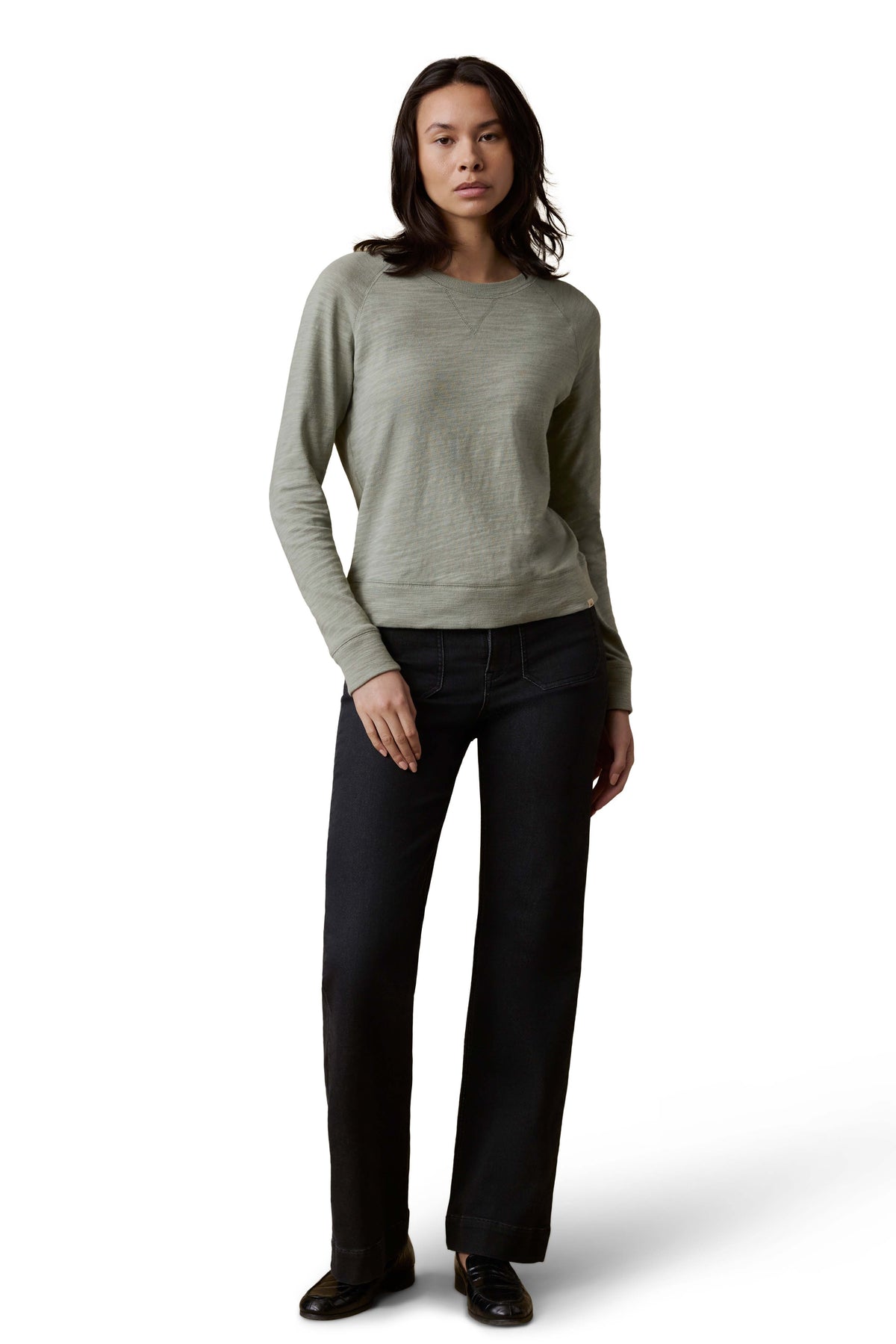 A woman wearing a sage green crewneck, dark jeans, and loafers, image 2 of 3