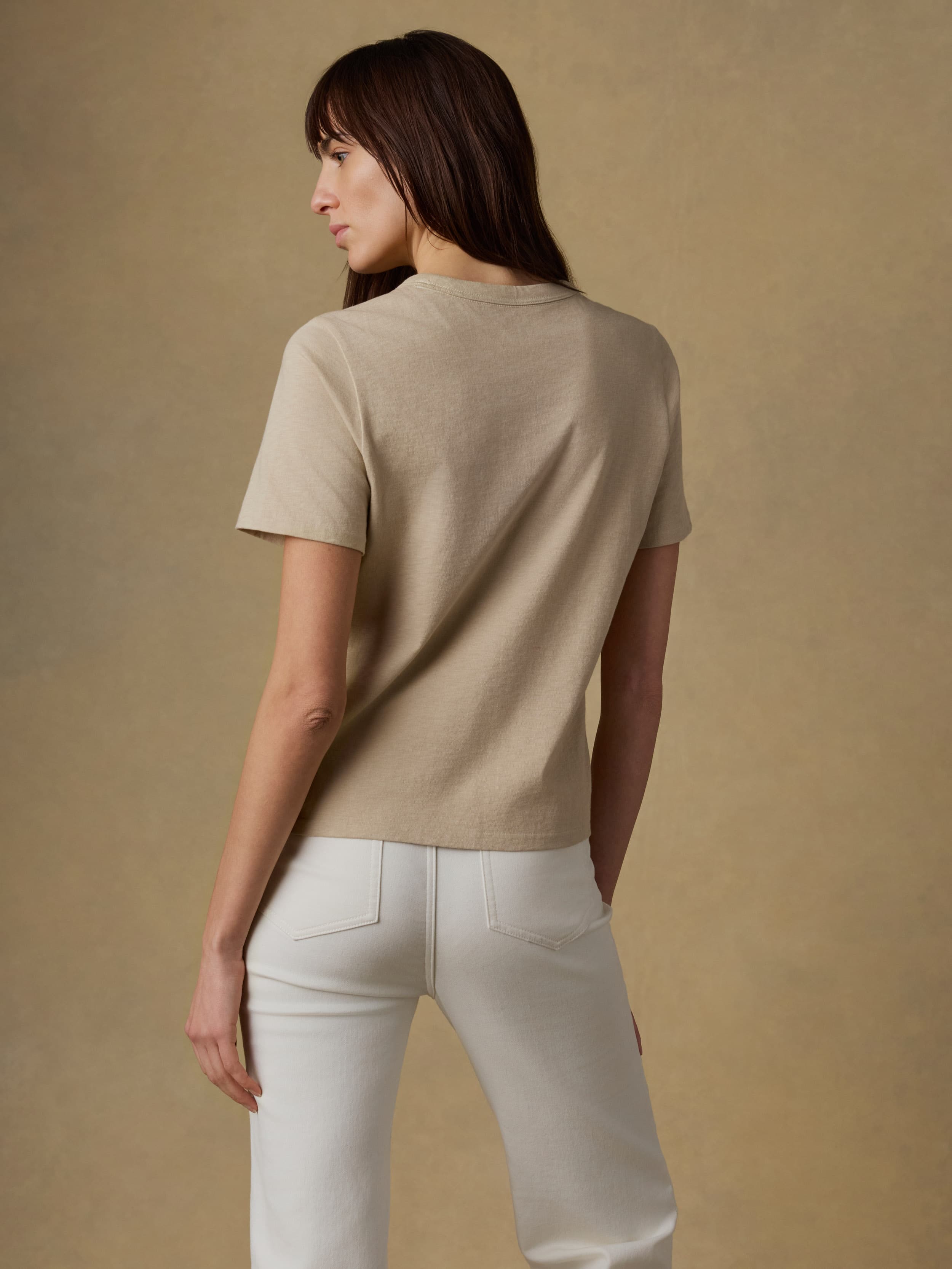 Back view of a woman wearing a beige t-shirt and white jeans against a beige background