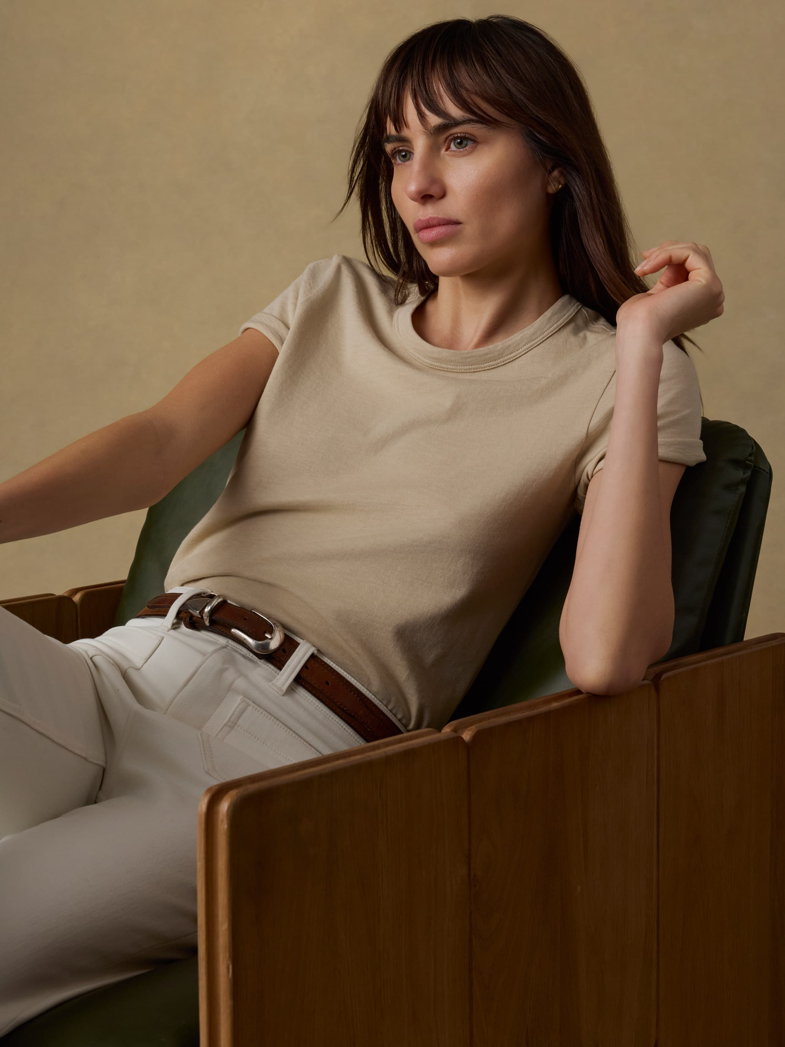 Woman sitting on a wooden chair wearing a beige t-shirt and white jeans against a beige background