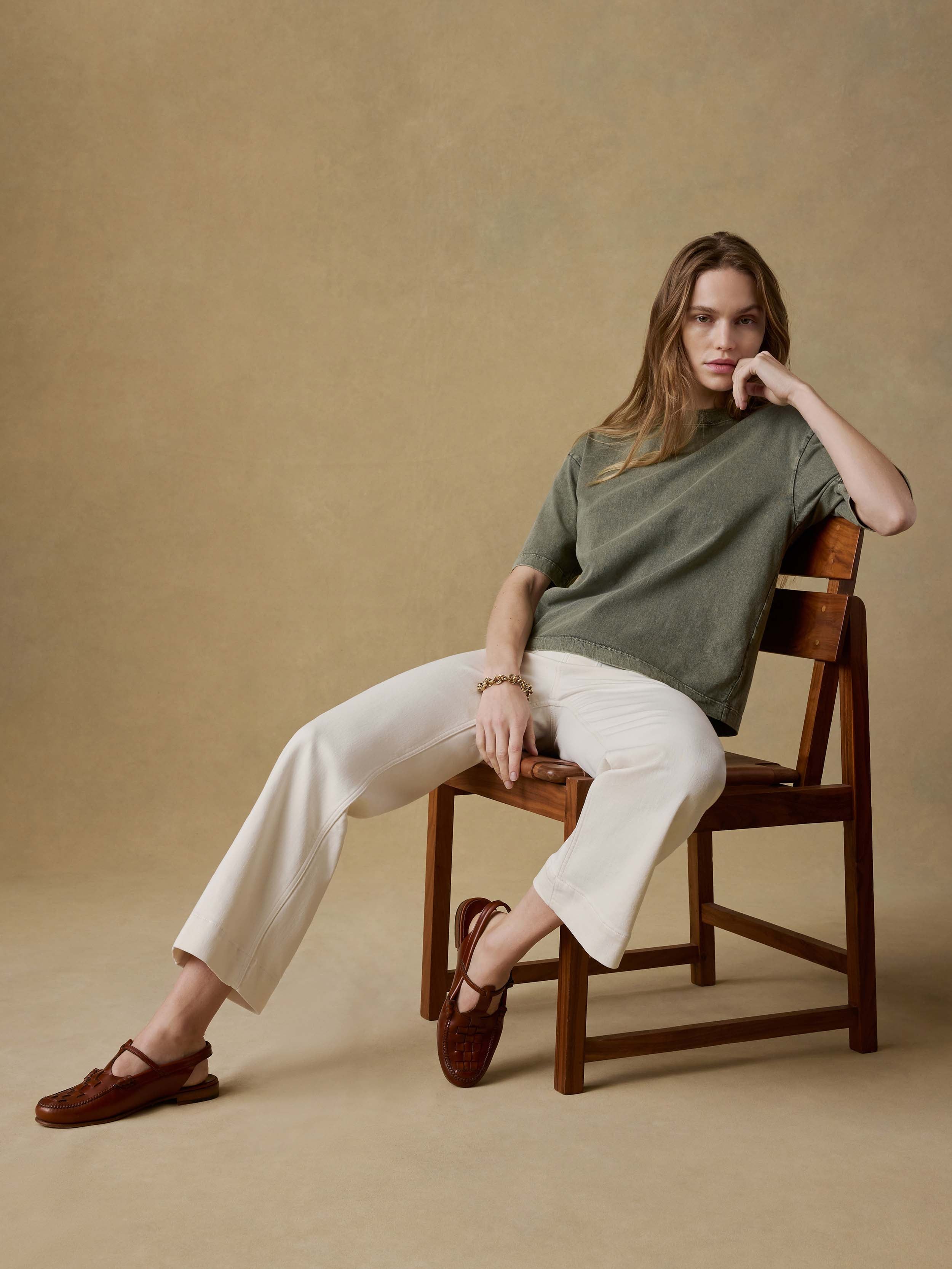 Woman sitting on a wooden chair wearing a green t-shirt and white pants against a beige background