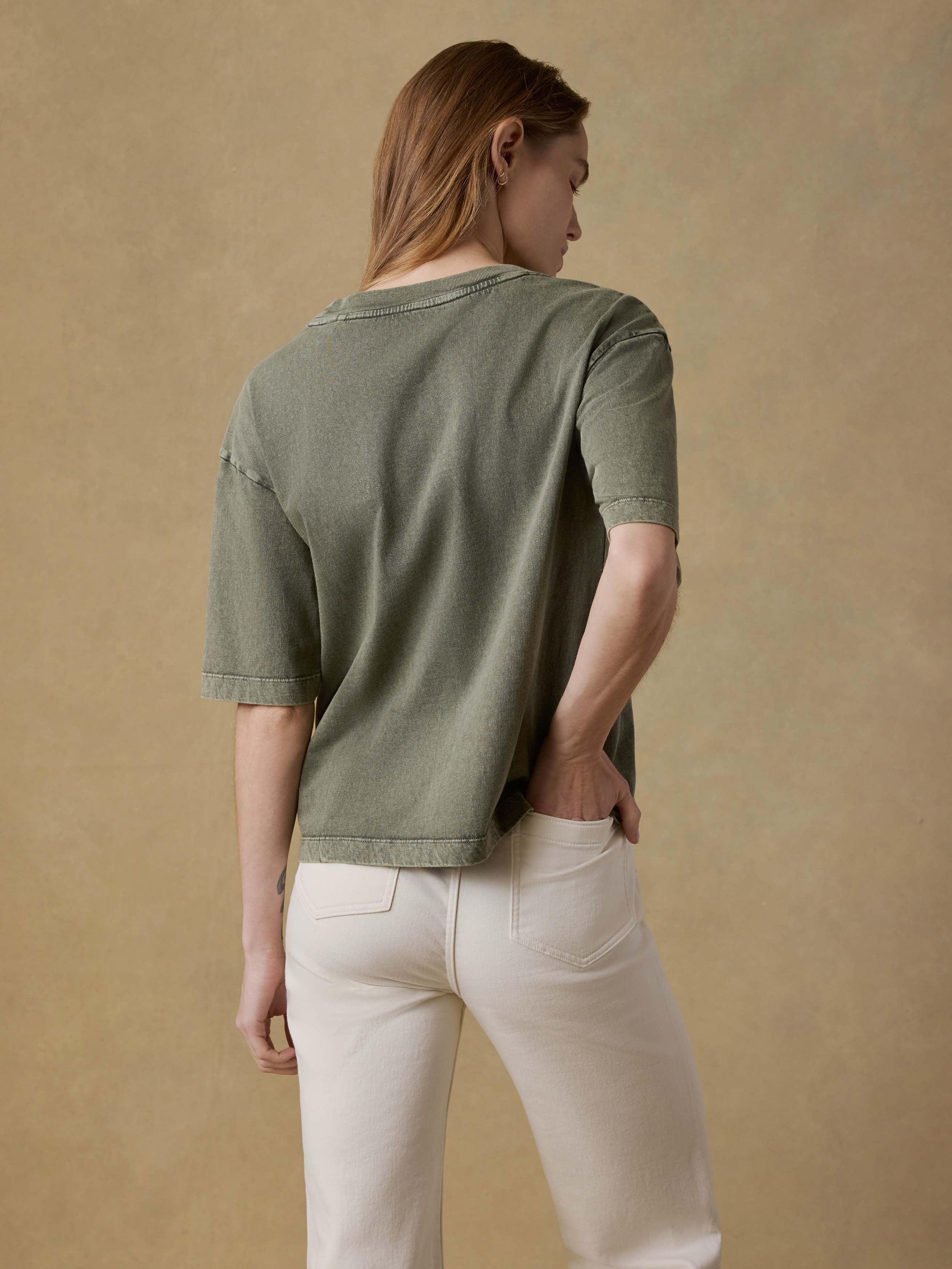 Back view of a woman wearing a green t-shirt and white pants against a beige background