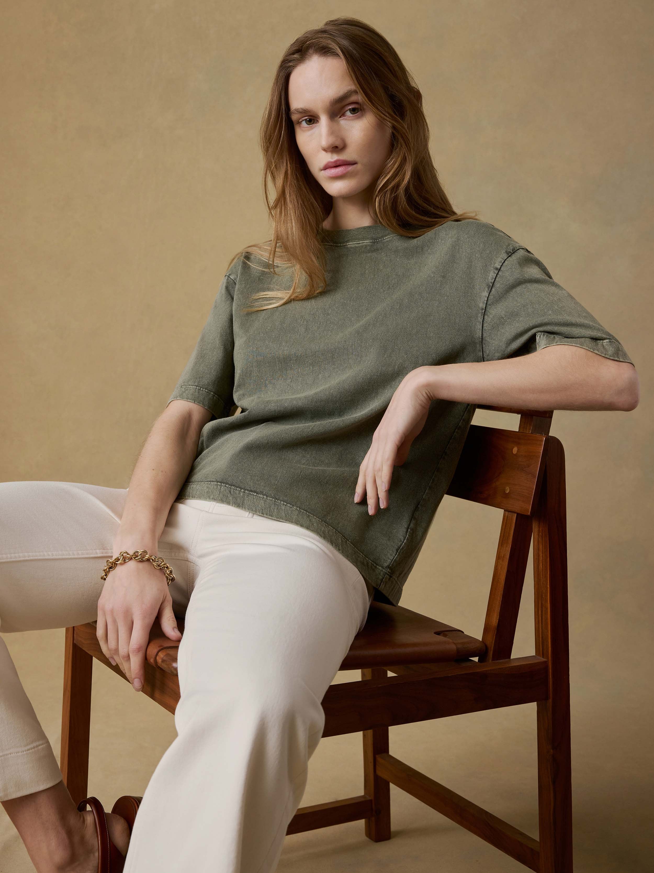 Woman sitting on a wooden chair wearing a green t-shirt and white pants against a beige background