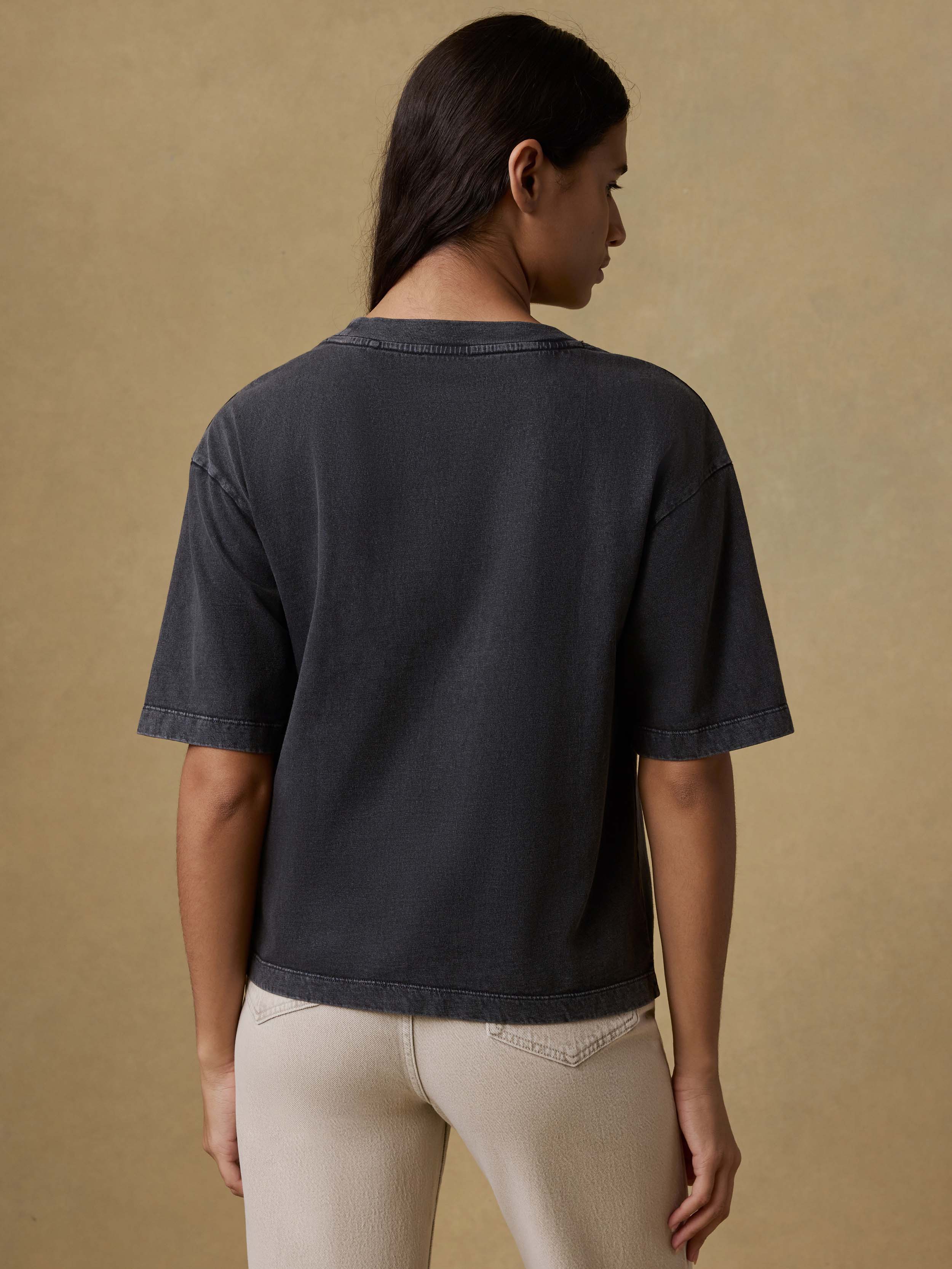 Back view of a woman wearing a black t-shirt and beige pants against a brown background