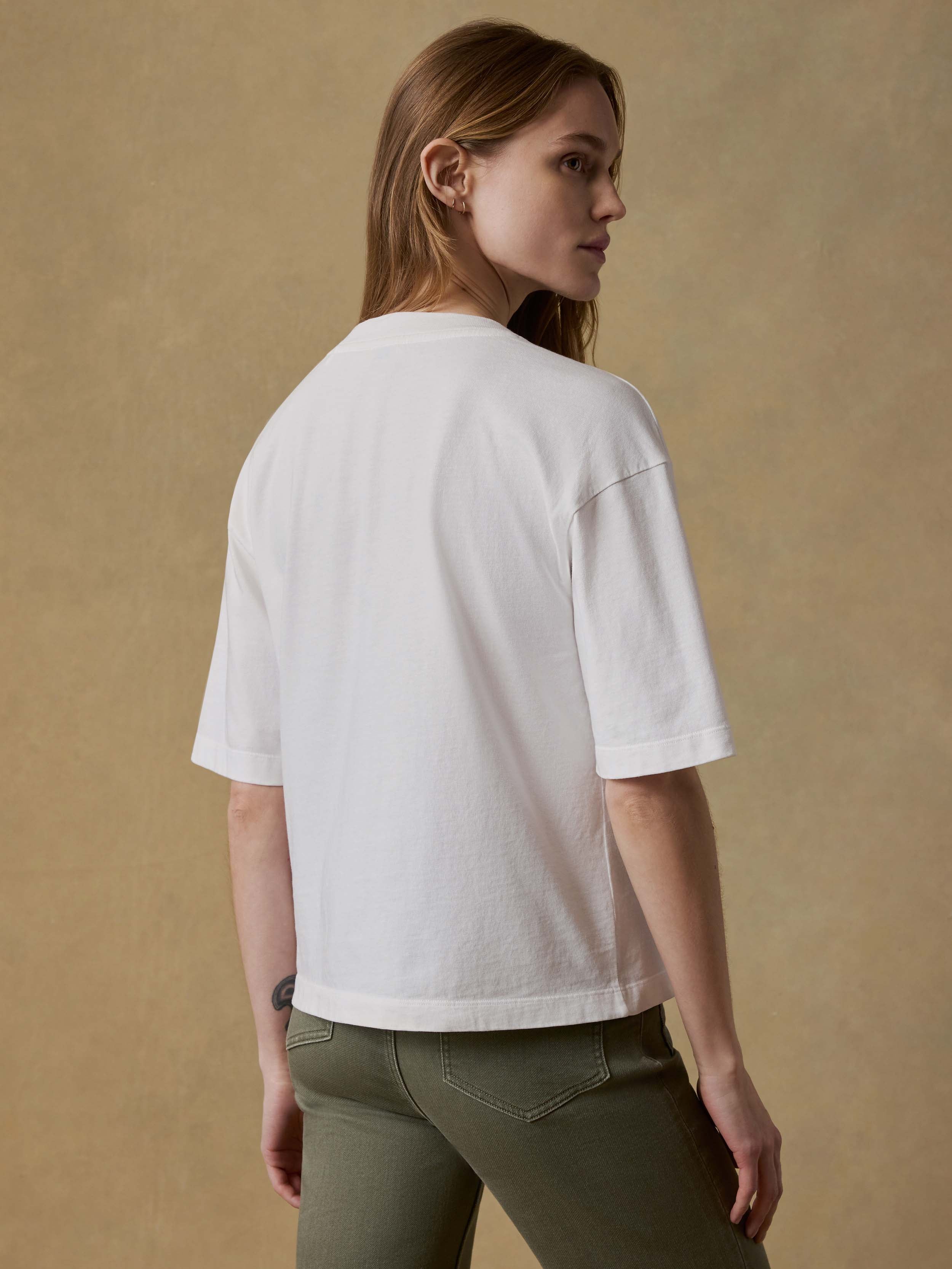 Back view of a woman wearing a white t-shirt and green pants against a beige background