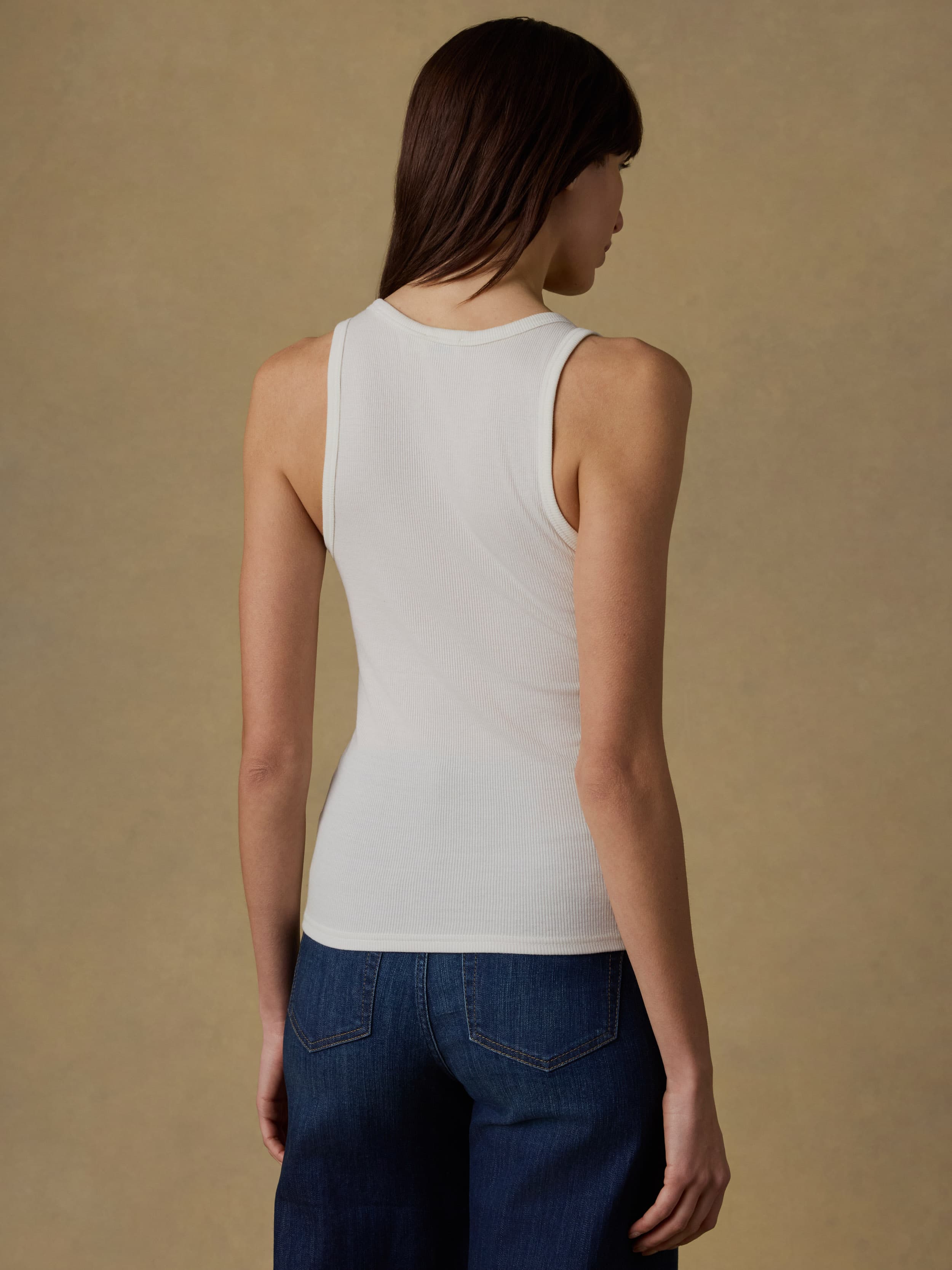 Back view of a woman wearing a white tank top and blue jeans against a beige background