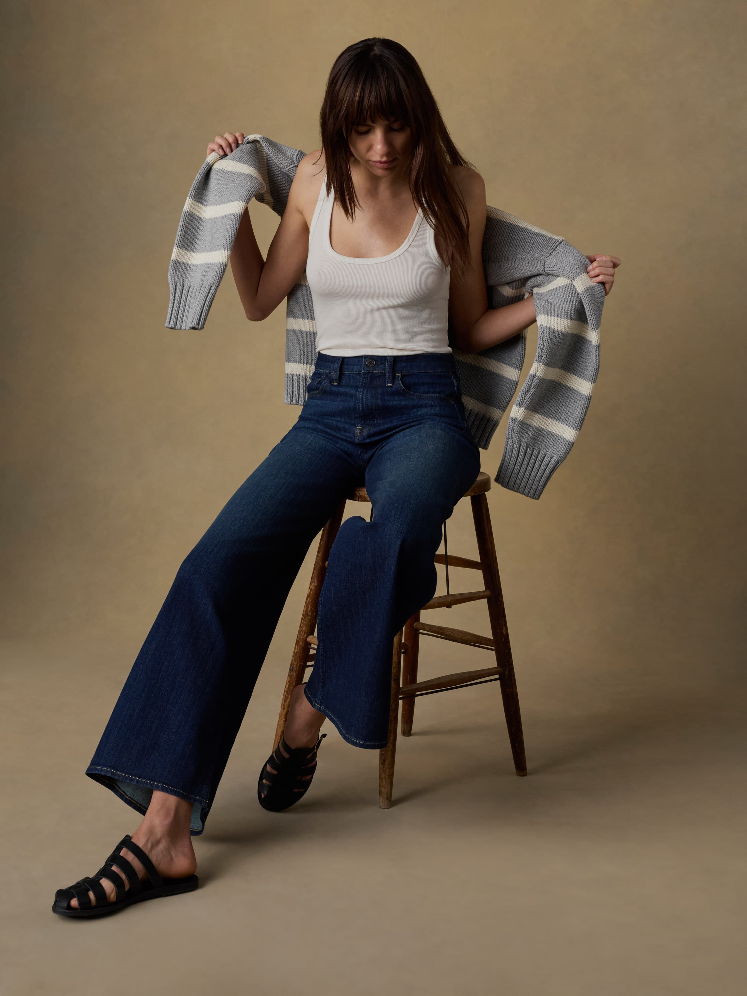 Woman sitting on a wooden stool wearing a white tank top and blue jeans against a beige background