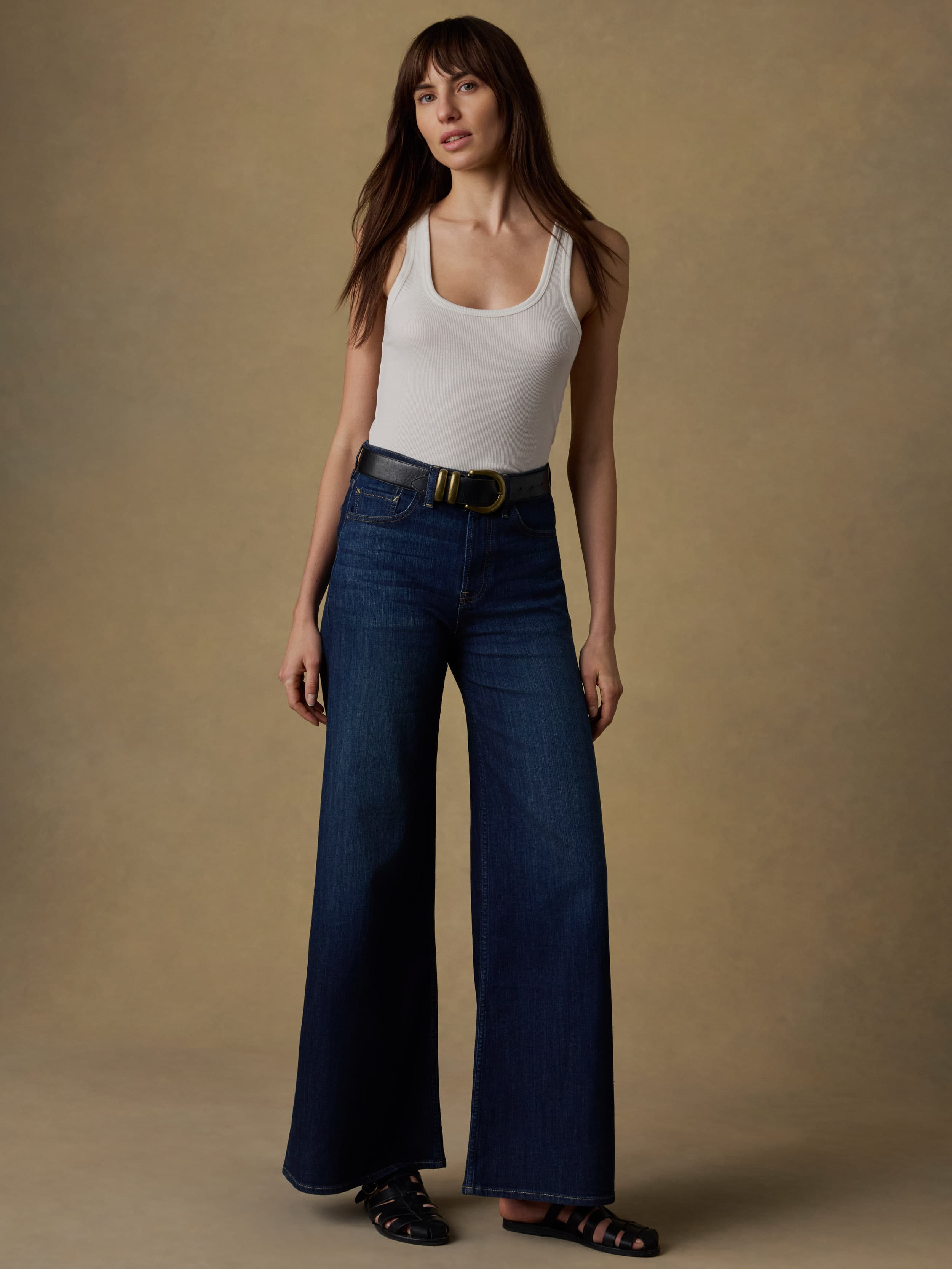 Woman wearing a white tank top and blue jeans against a beige background