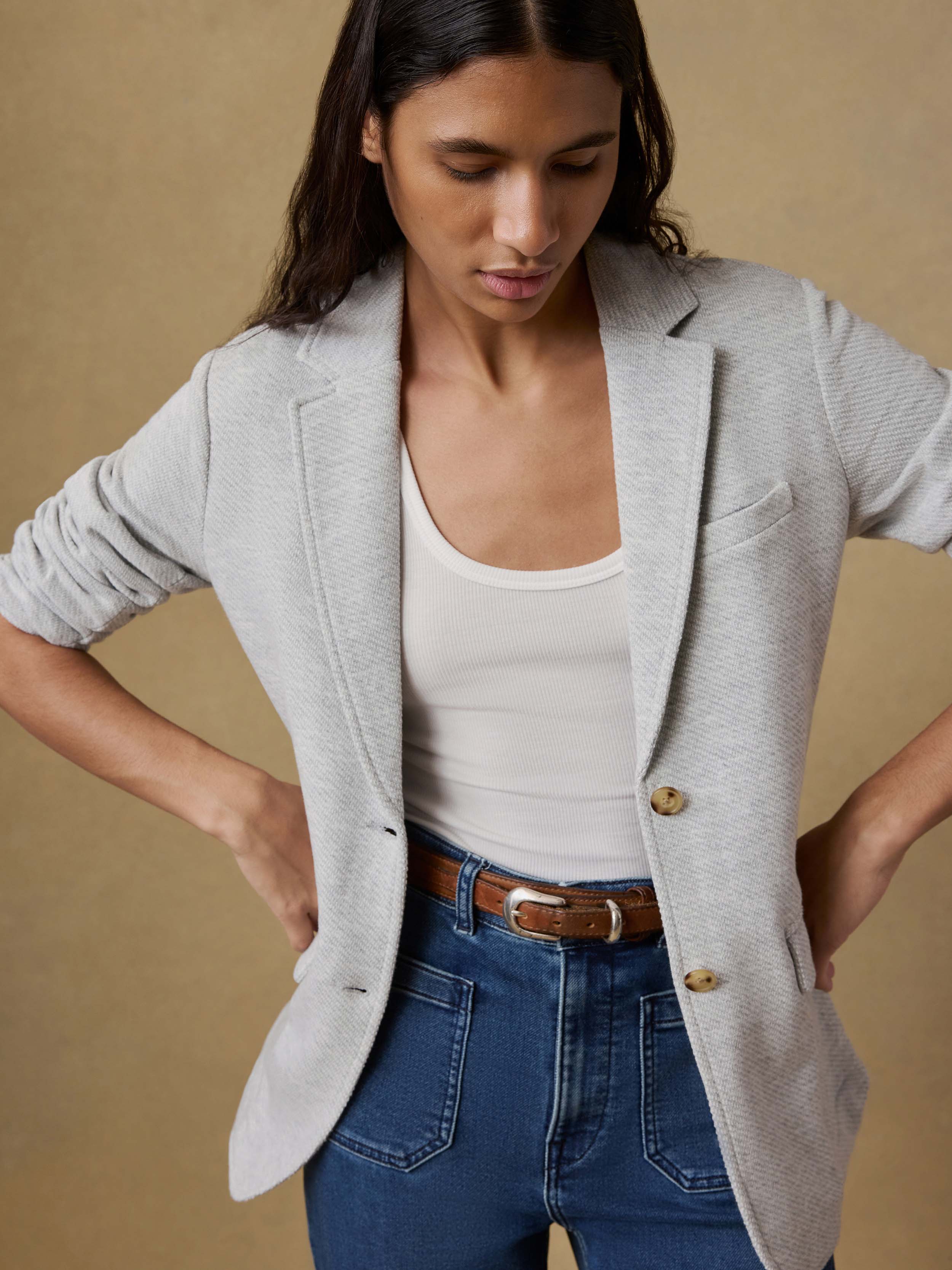 Woman wearing a light gray blazer over a white tank top and blue jeans on a beige background
