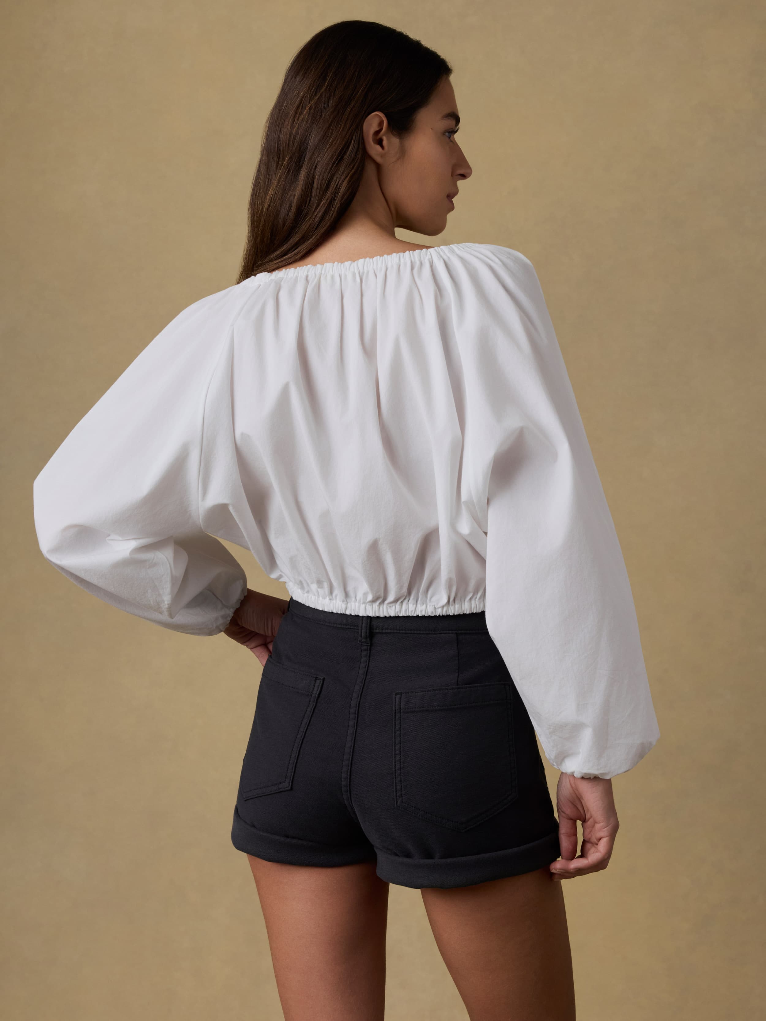 Back view of a woman wearing a white blouse with puffed sleeves and black shorts on a beige background