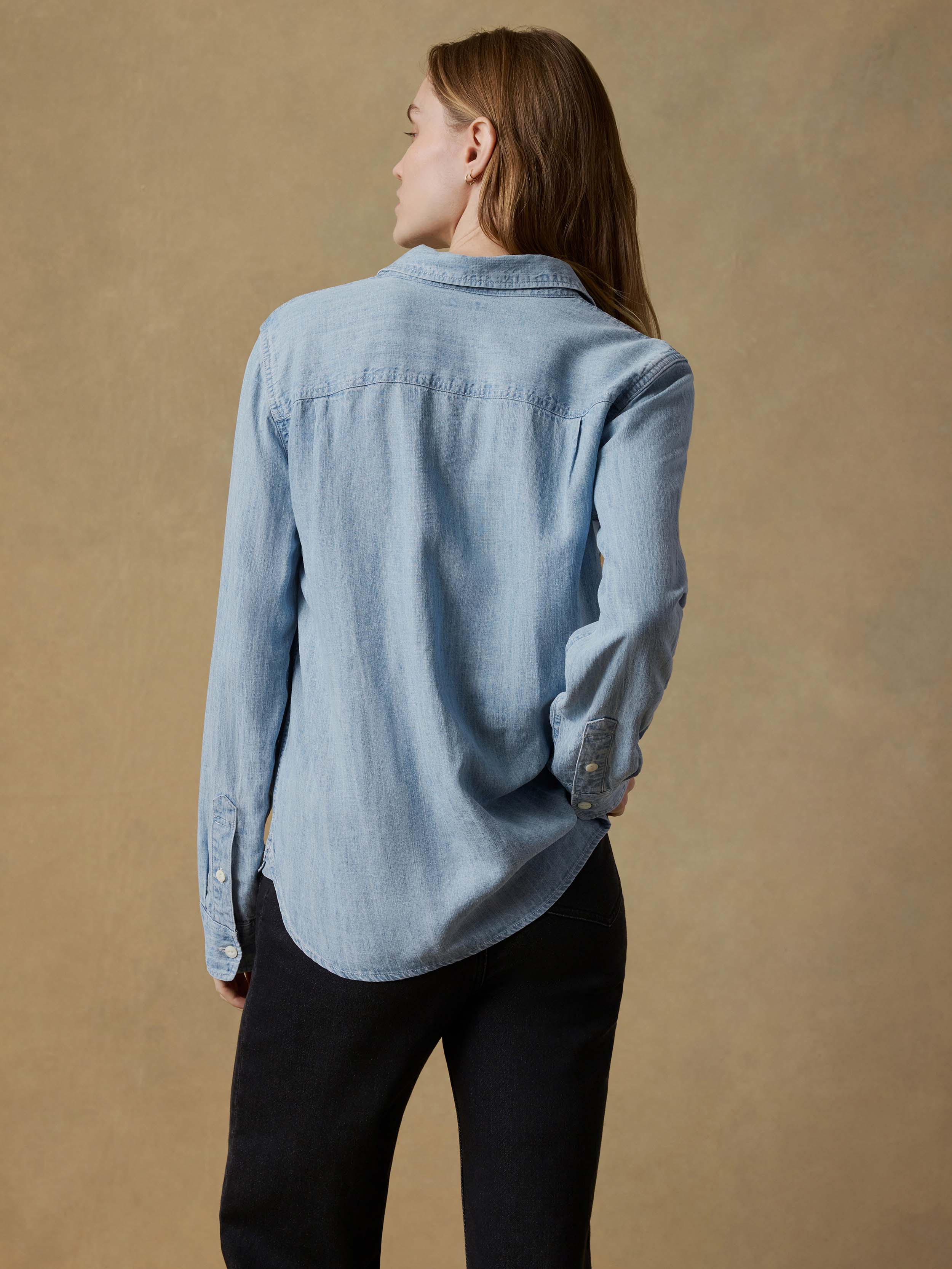 Back view of a woman wearing a light blue shirt and black jeans against a beige background