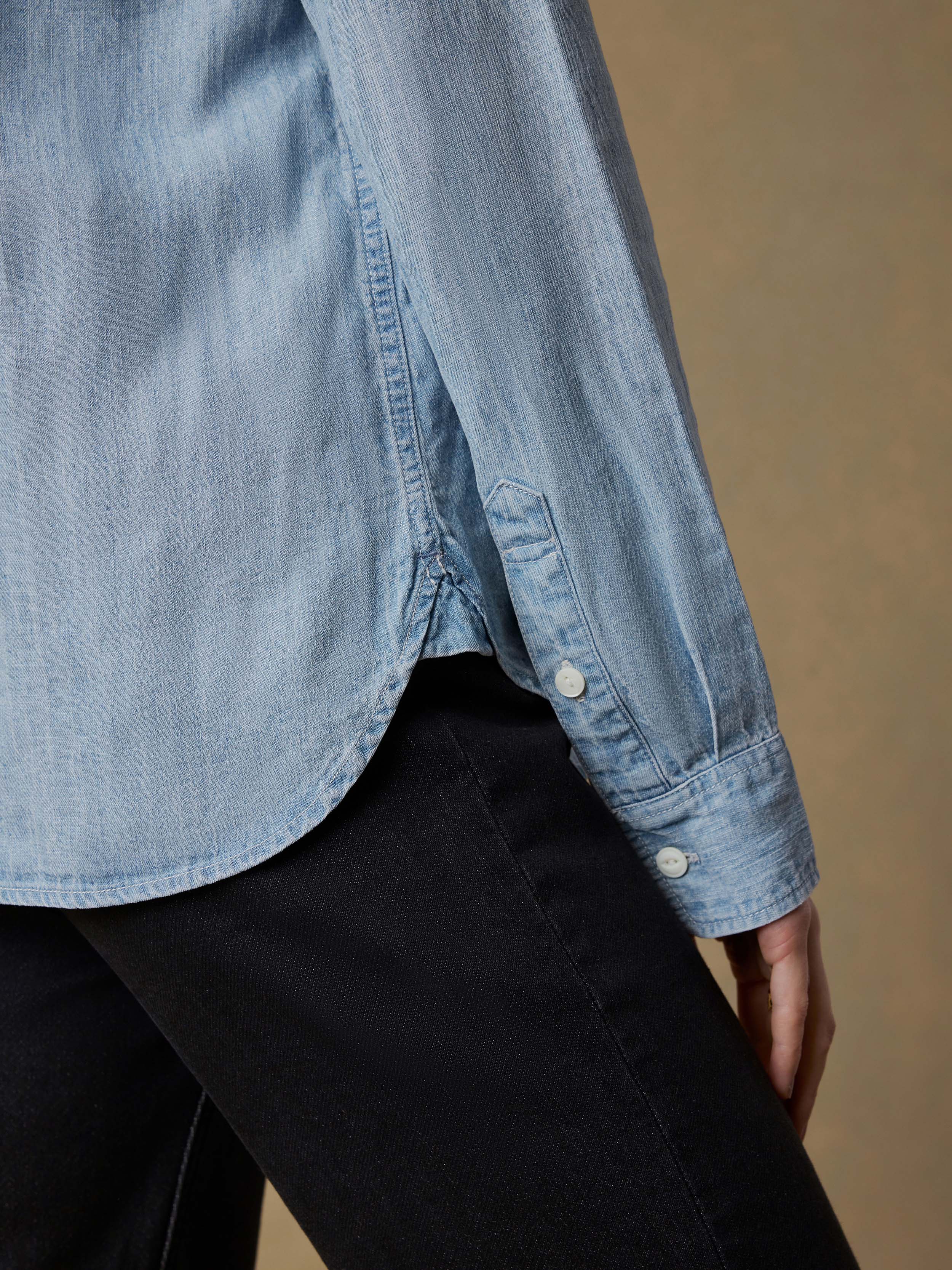 Detail shot of a woman wearing a light blue denim shirt with black jeans against a beige background