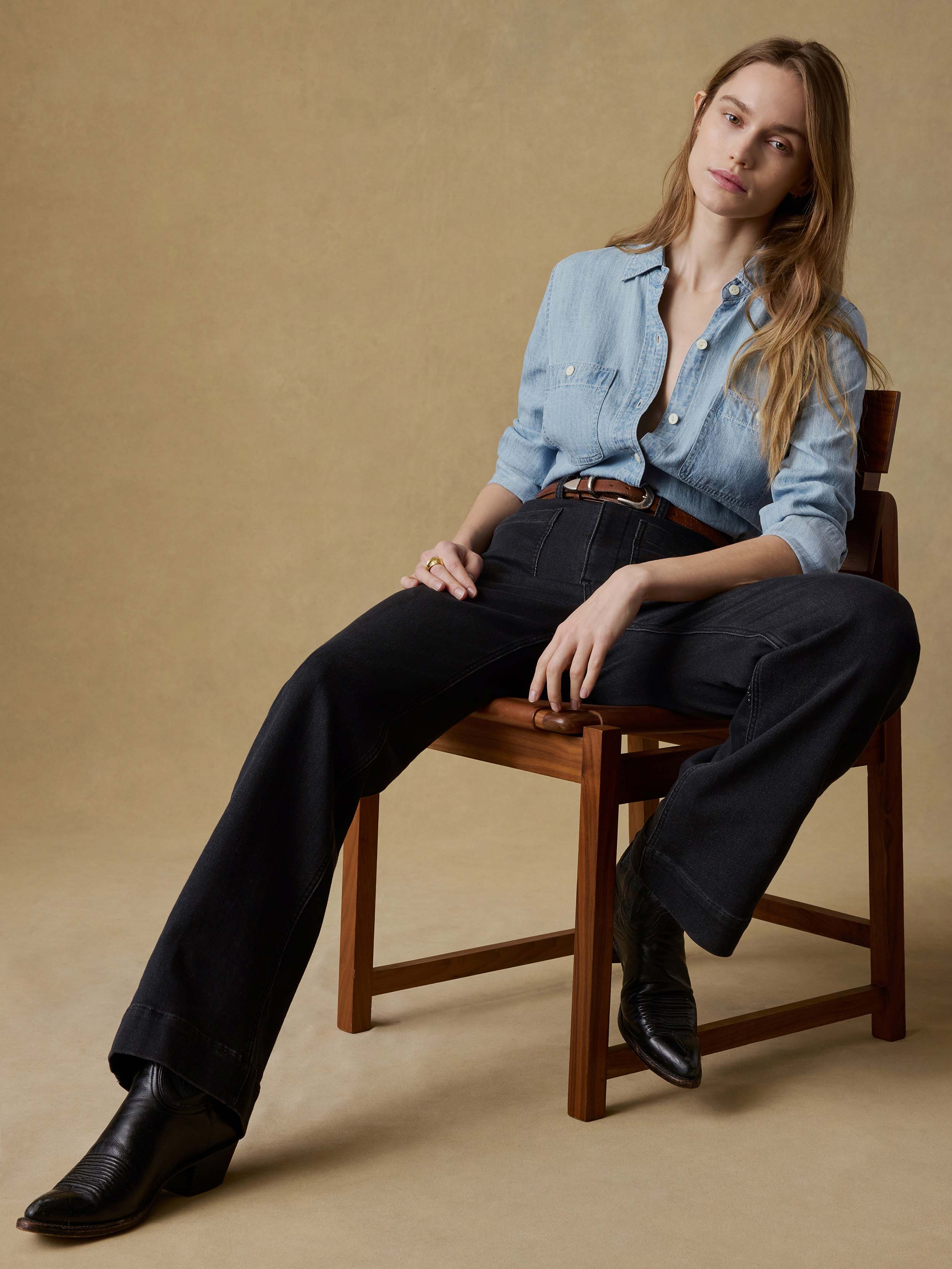 Woman sitting on a wooden chair wearing a light blue shirt and black jeans against a beige background