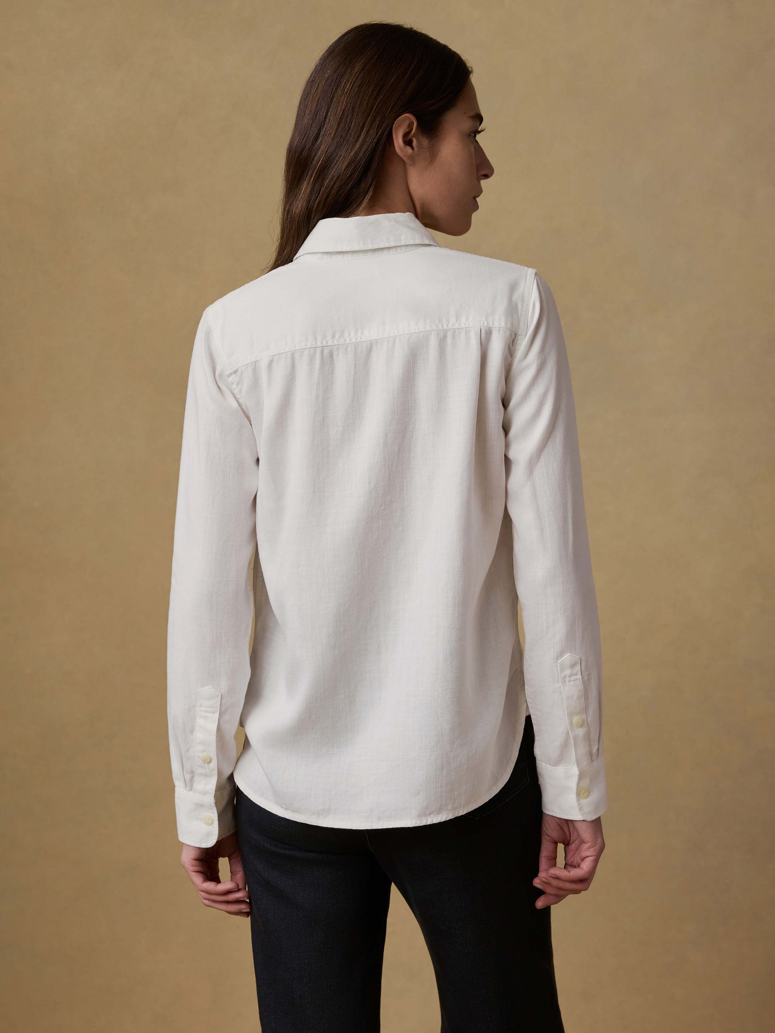Back view of a woman wearing a white chambray shirt and black jeans against a beige background