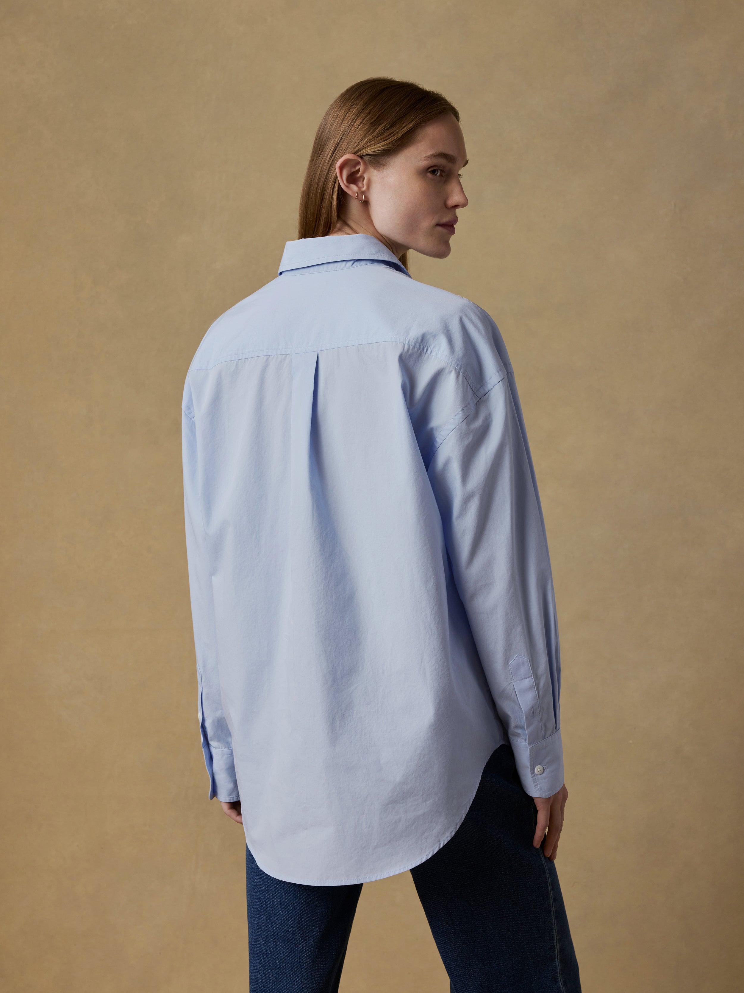 Back view of a woman wearing a light blue shirt and blue jeans against a beige background