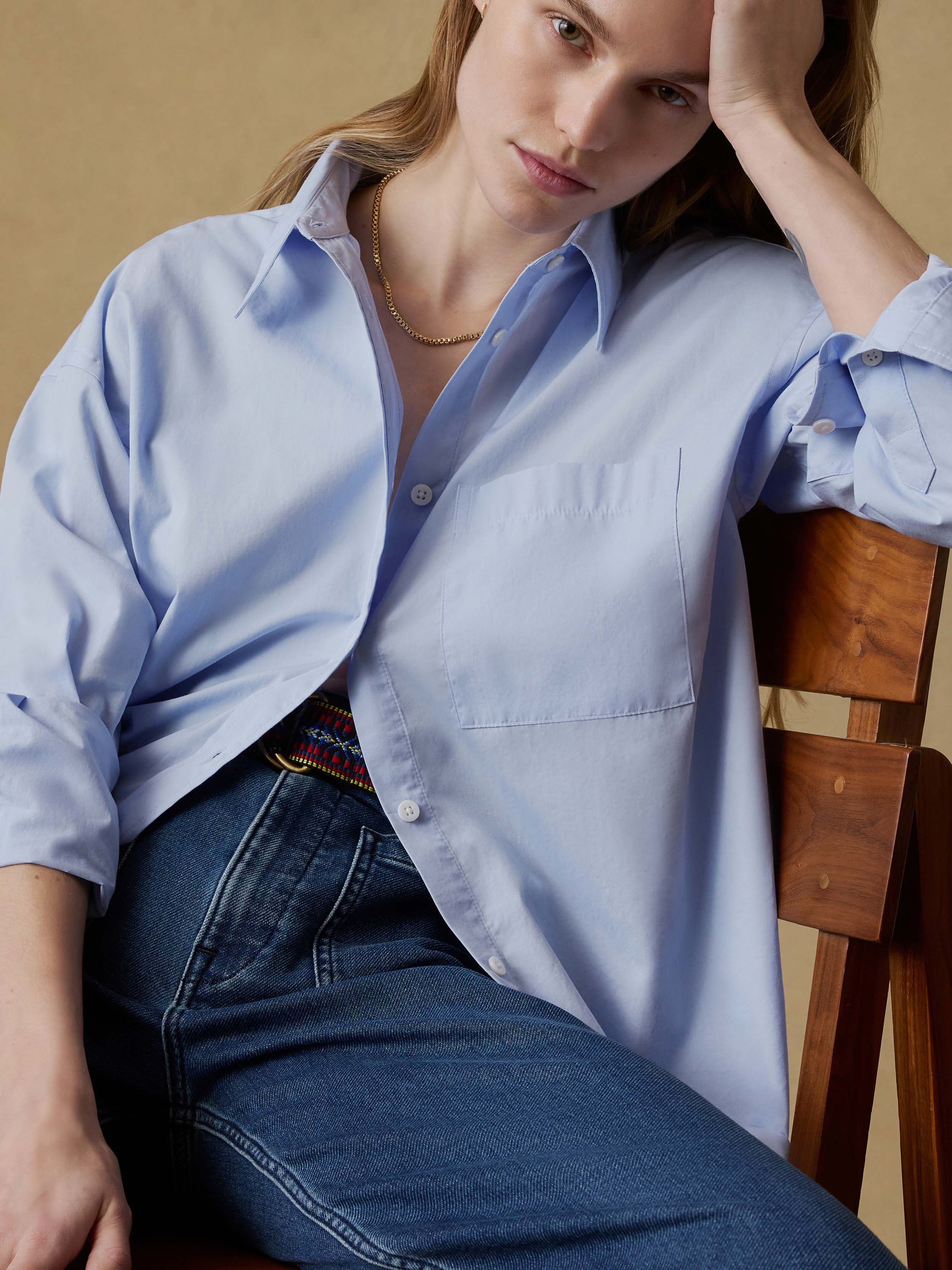 Woman sitting on a wooden chair wearing a light blue shirt and blue jeans against a beige background