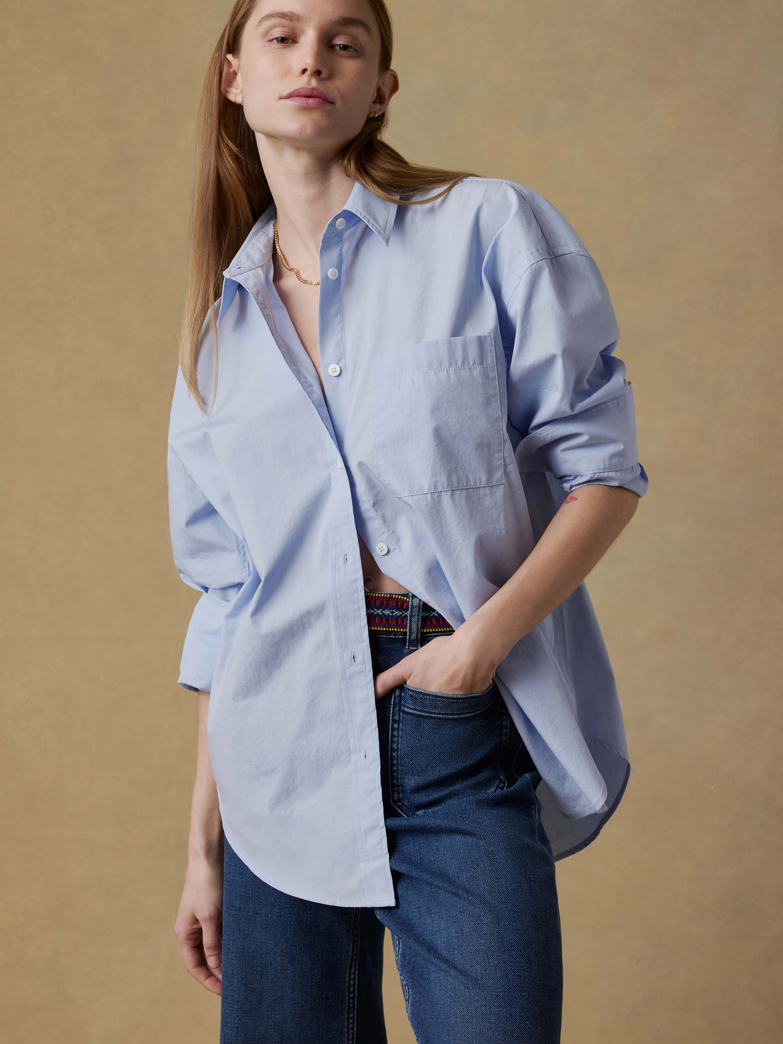 Woman wearing a light blue oversized shirt and dark jeans against a beige background