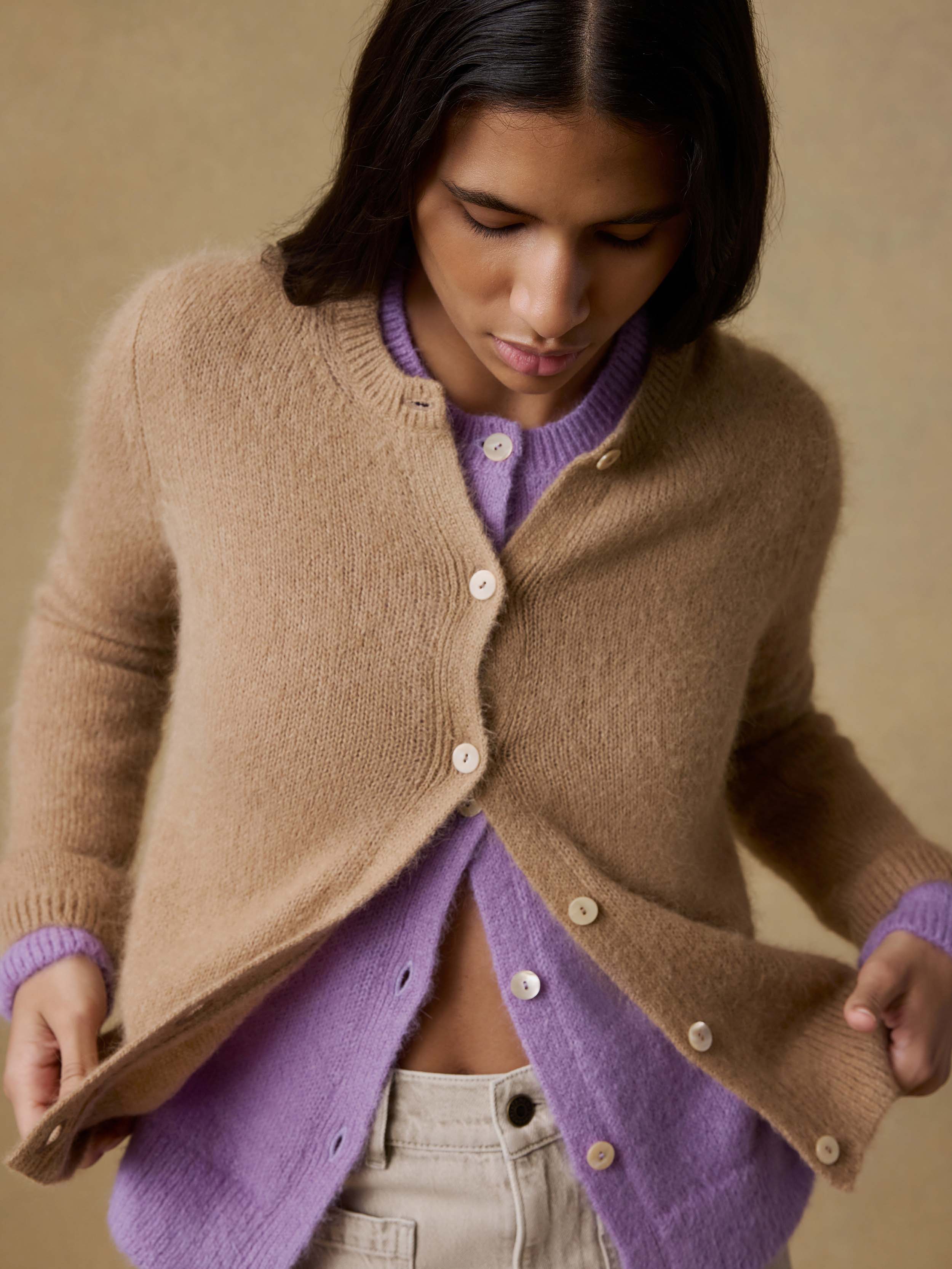 Woman wearing a beige cardigan over a purple cardigan with beige pants against a beige background