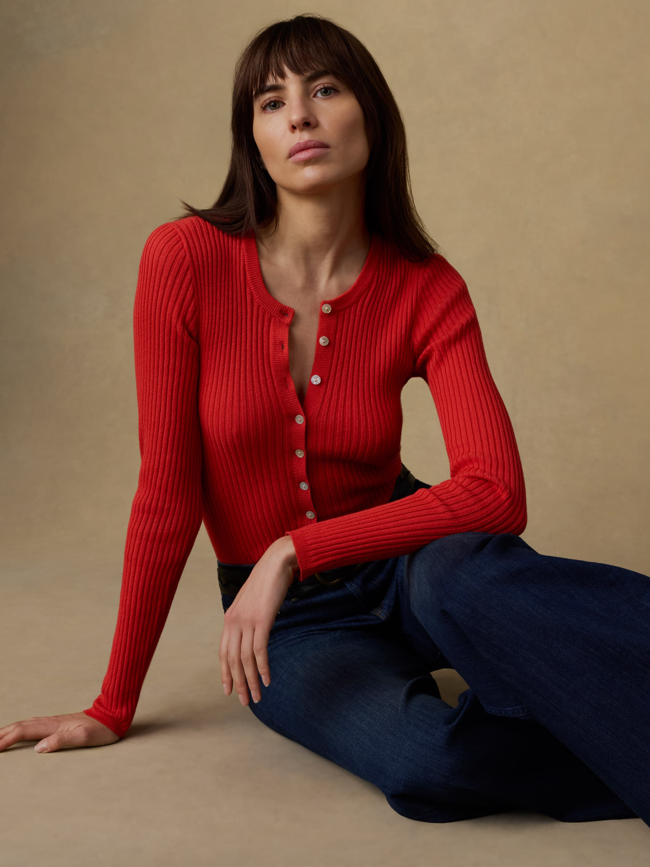 Woman wearing a red ribbed sweater and blue jeans against a beige background