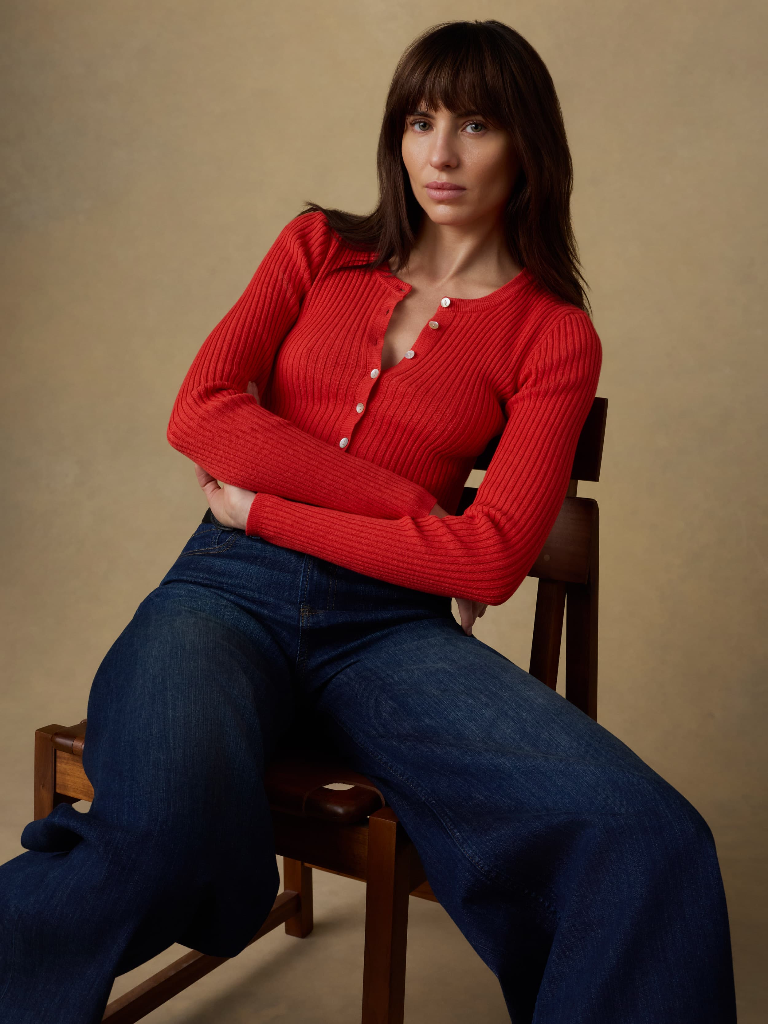 Woman sitting on a wooden chair wearing a red ribbed cardigan and blue jeans against a beige background