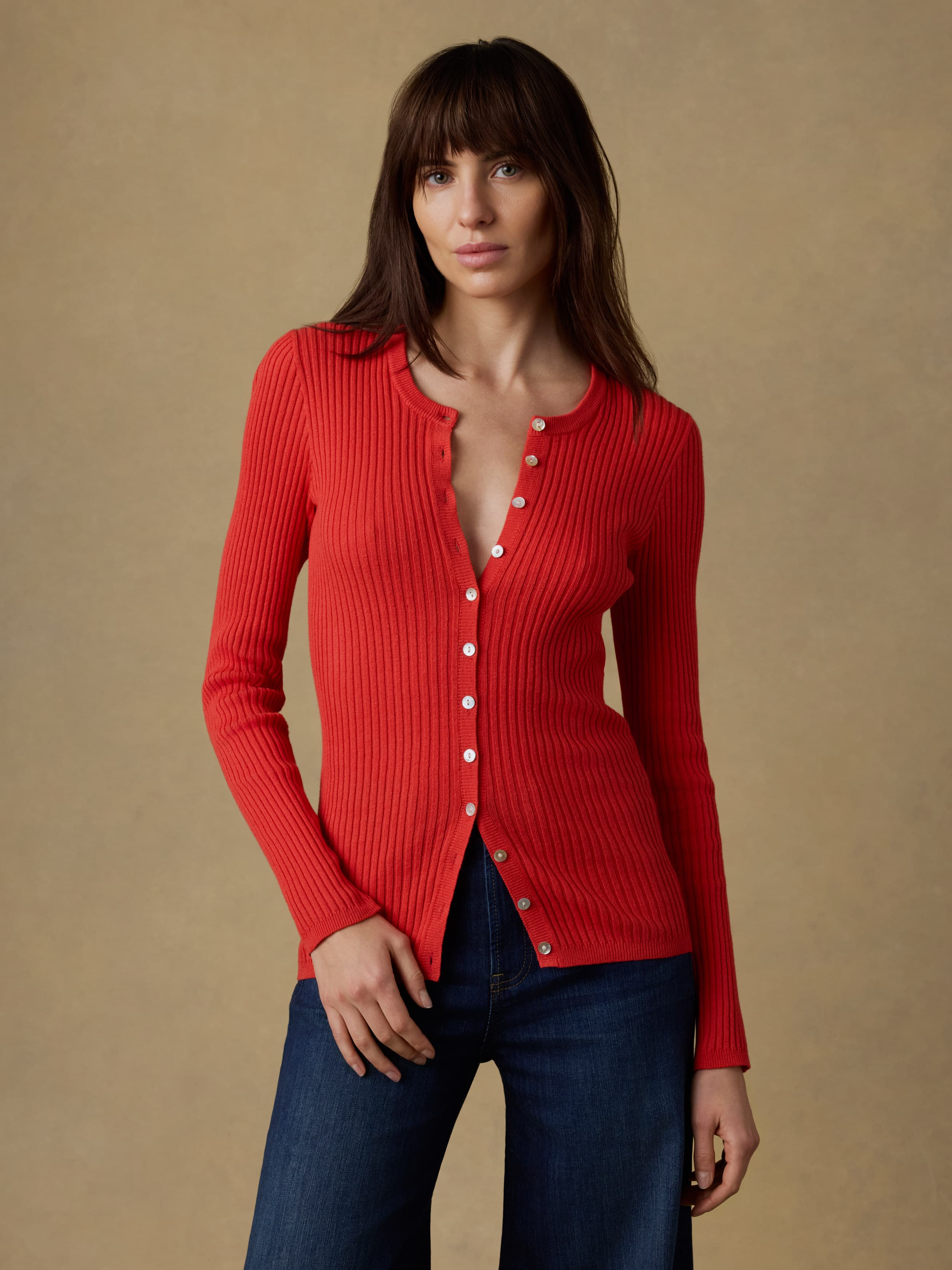 Woman wearing a red ribbed cardigan and blue jeans against a beige background