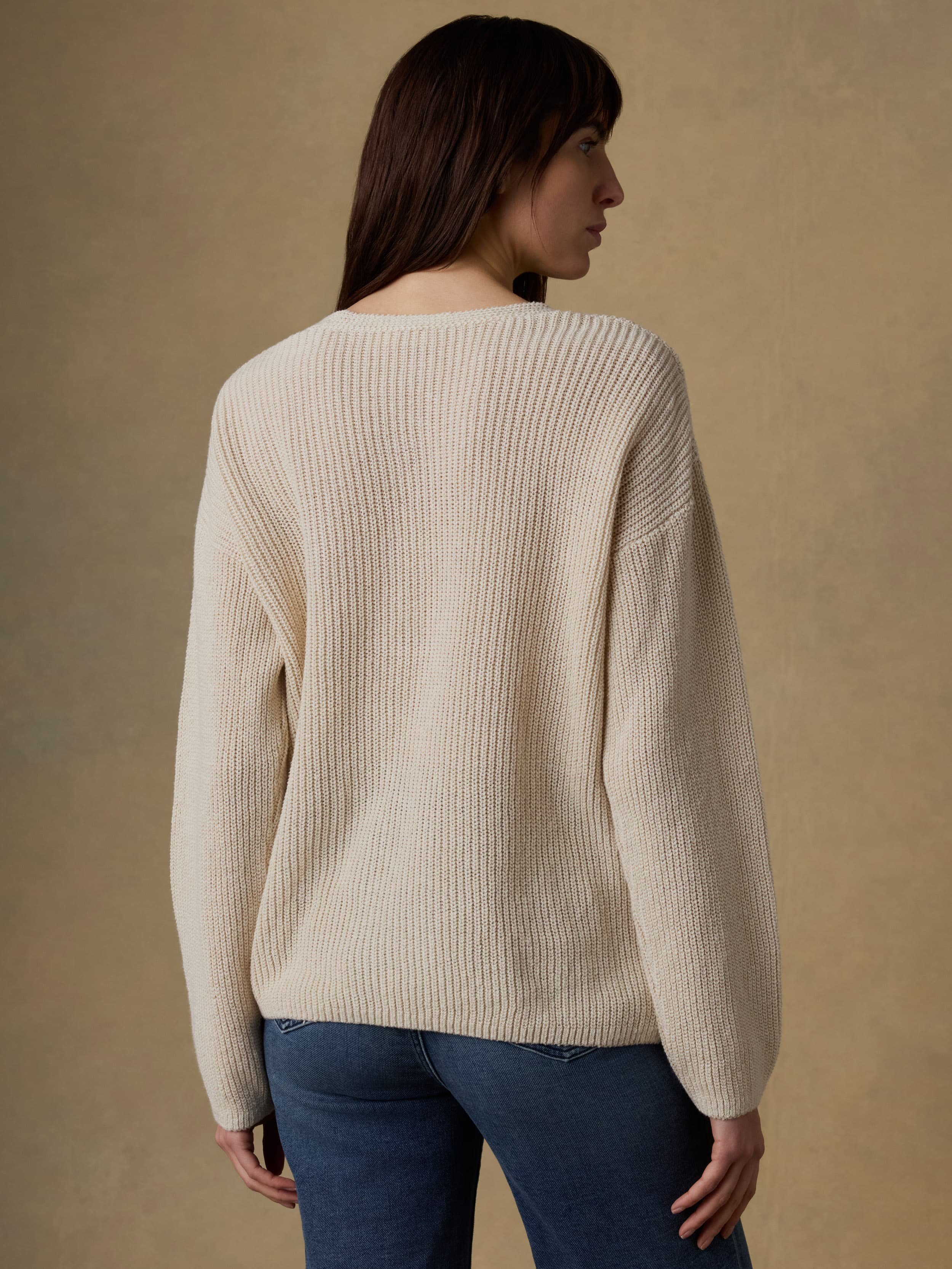 Back view of a woman wearing a beige sweater and blue jeans against a beige background