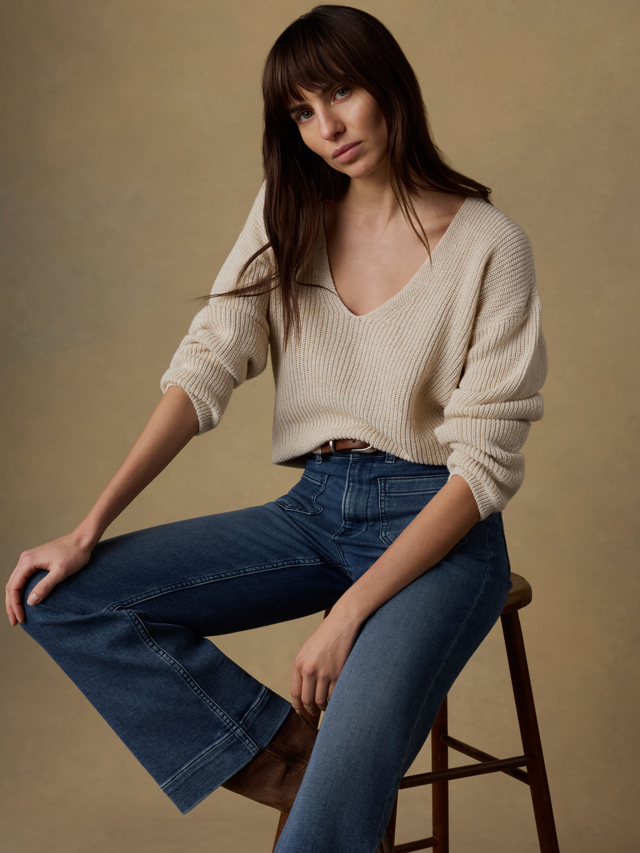 Woman wearing a beige sweater and blue jeans sitting on a wooden stool against a beige background