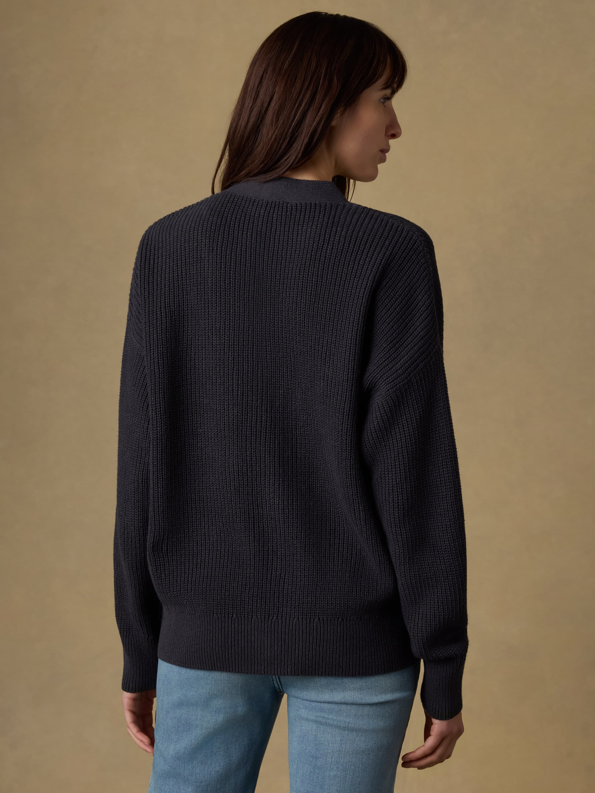 Back view of a woman wearing a black knit cardigan and light blue jeans against a beige background