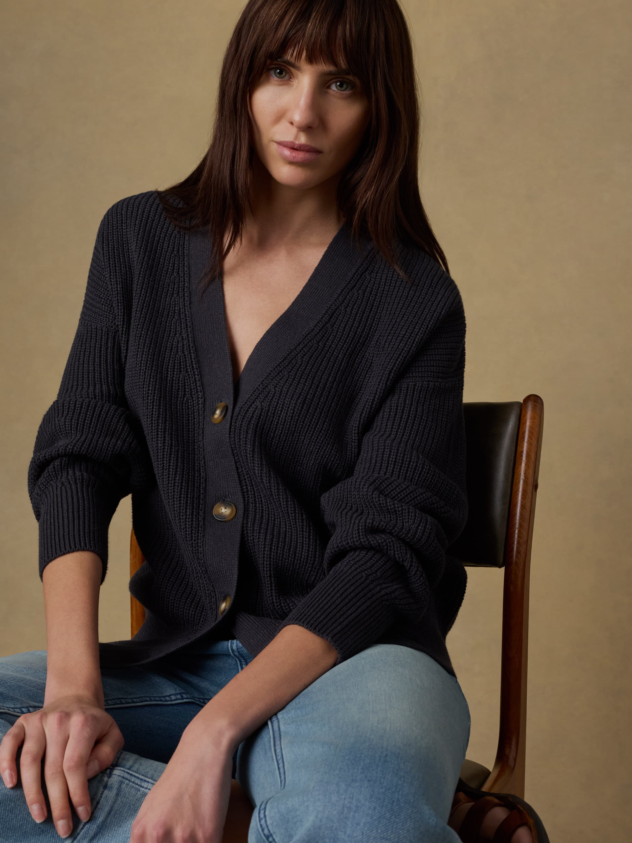 Woman sitting on a wooden chair wearing a black knit cardigan and light blue jeans against a beige background