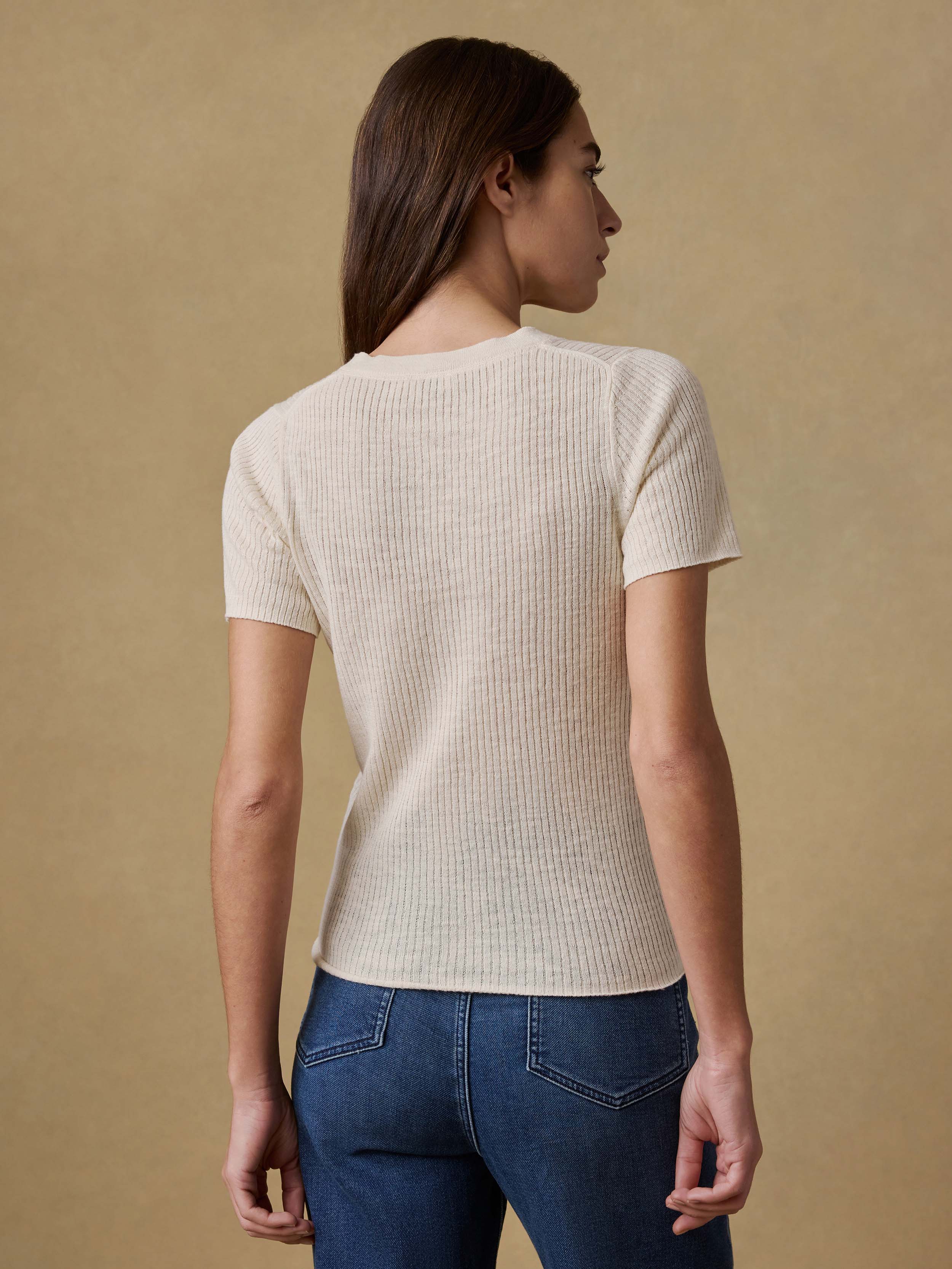Back view of a woman wearing a cream ribbed t-shirt and blue jeans against a beige background