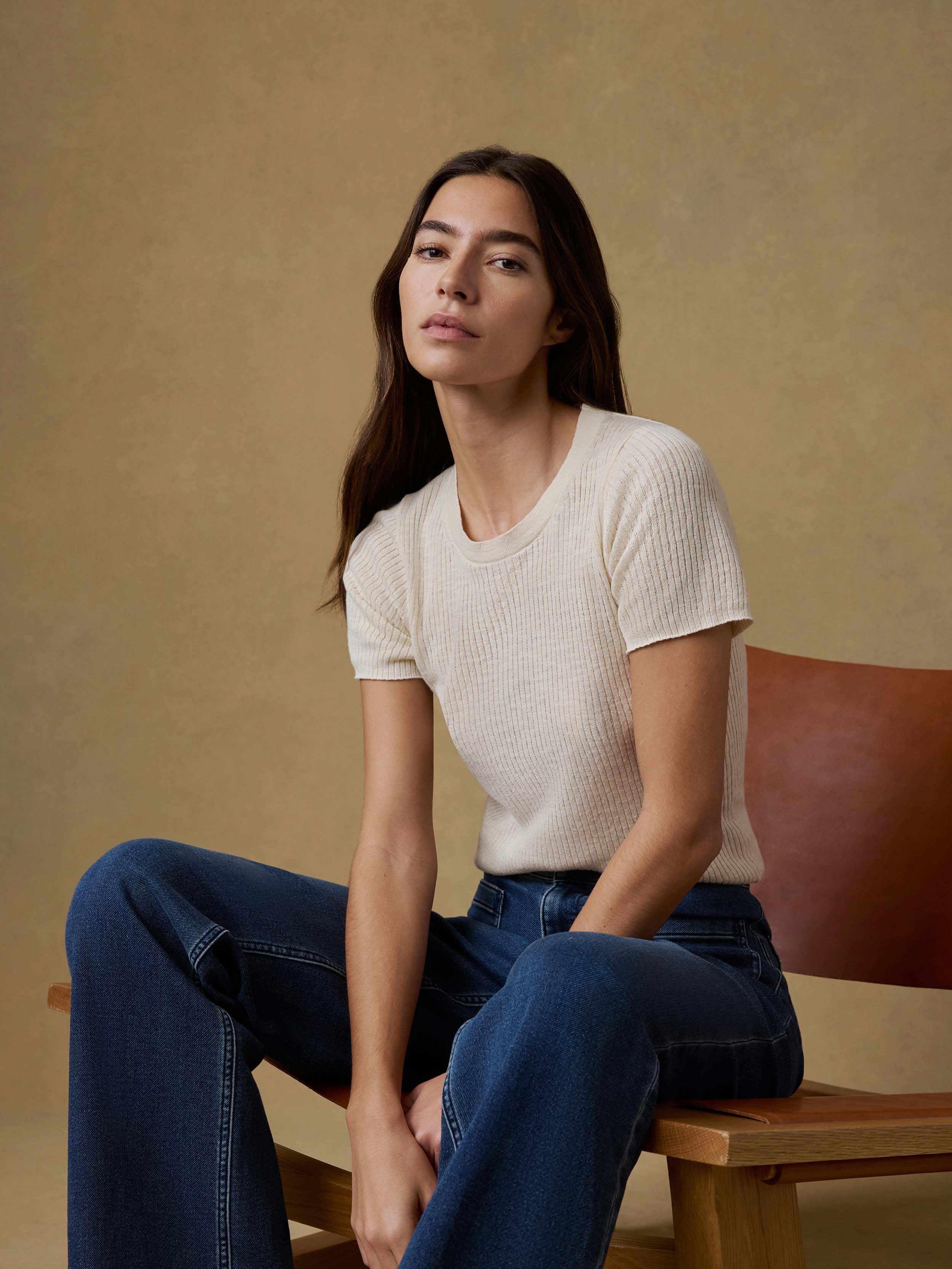 Woman sitting on a wooden chair wearing a white t-shirt and blue jeans against a beige background