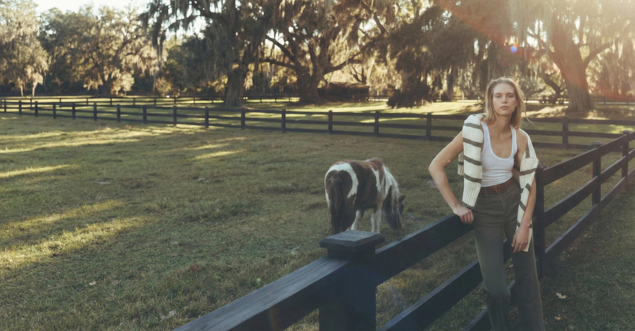 Female model leaning on a wooden fence with Charleston farm in background, a pony with braided hair in the background. She wears a tank top and green stretch terry patch pocket pants, wrapping her arms in a striped sweater.