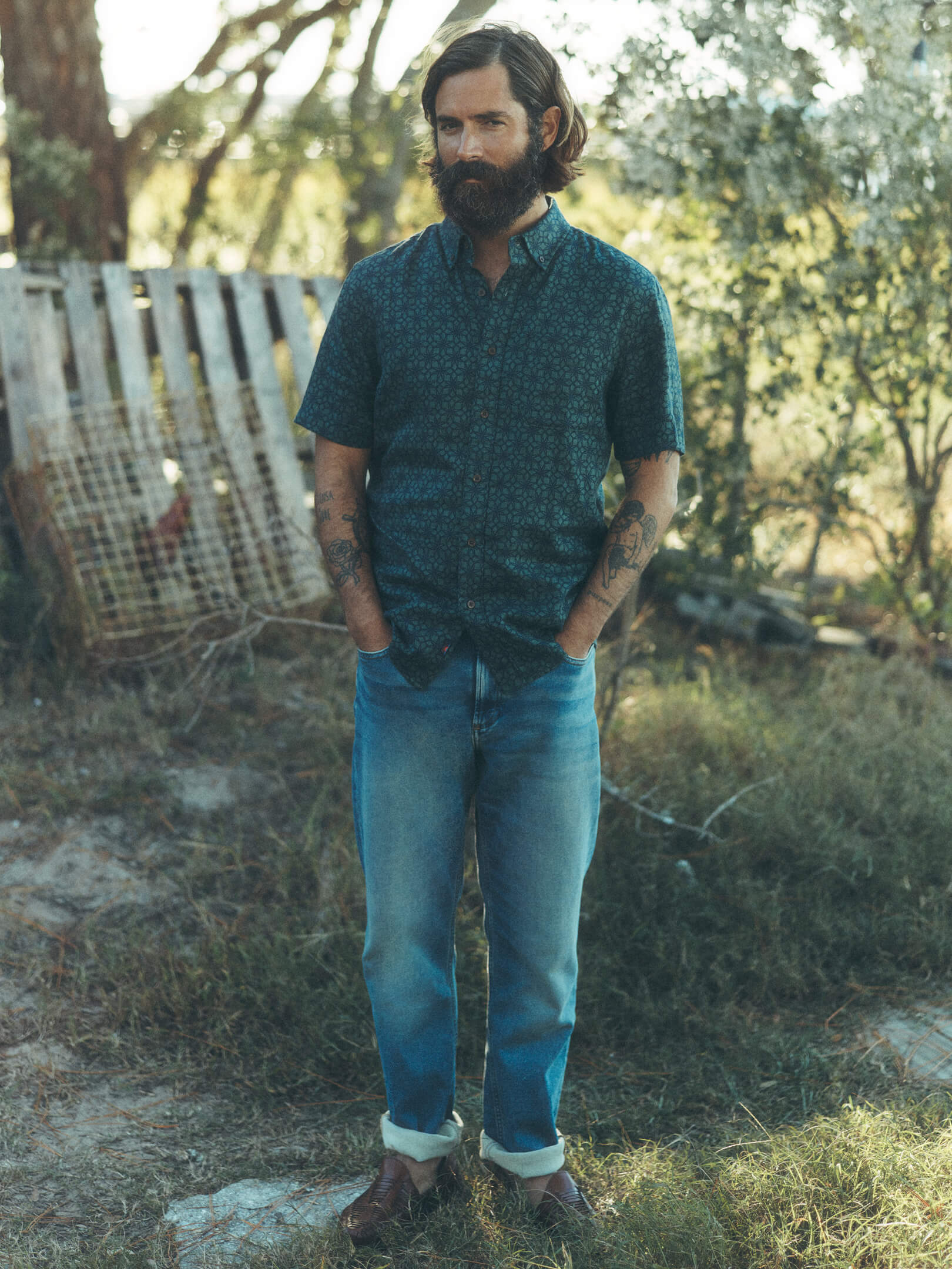 A male model in a southern garden, wearing stretch terry pants, huarache sandals and a short sleeve button up shirt.